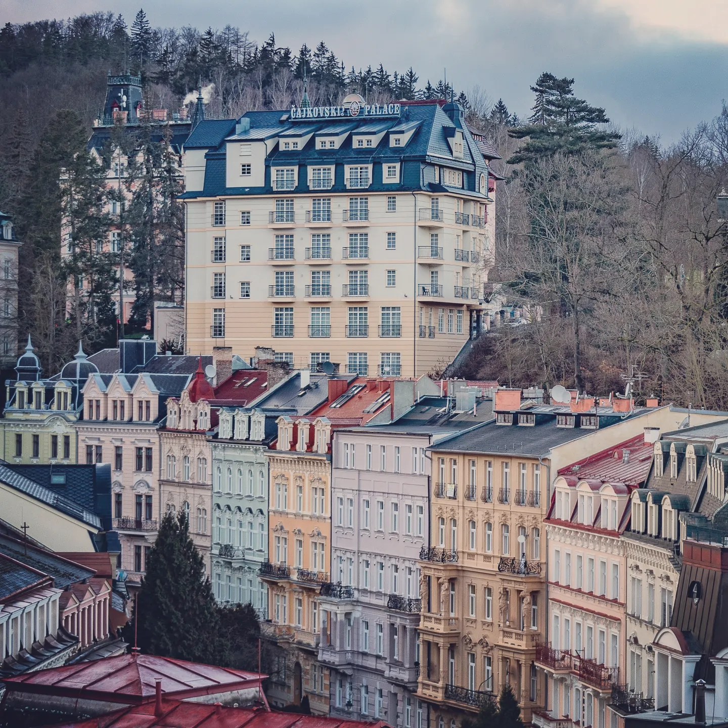 A row of pastel historic buildings lines a street below the large Cajkovskij Palace hotel set against wooded hills.