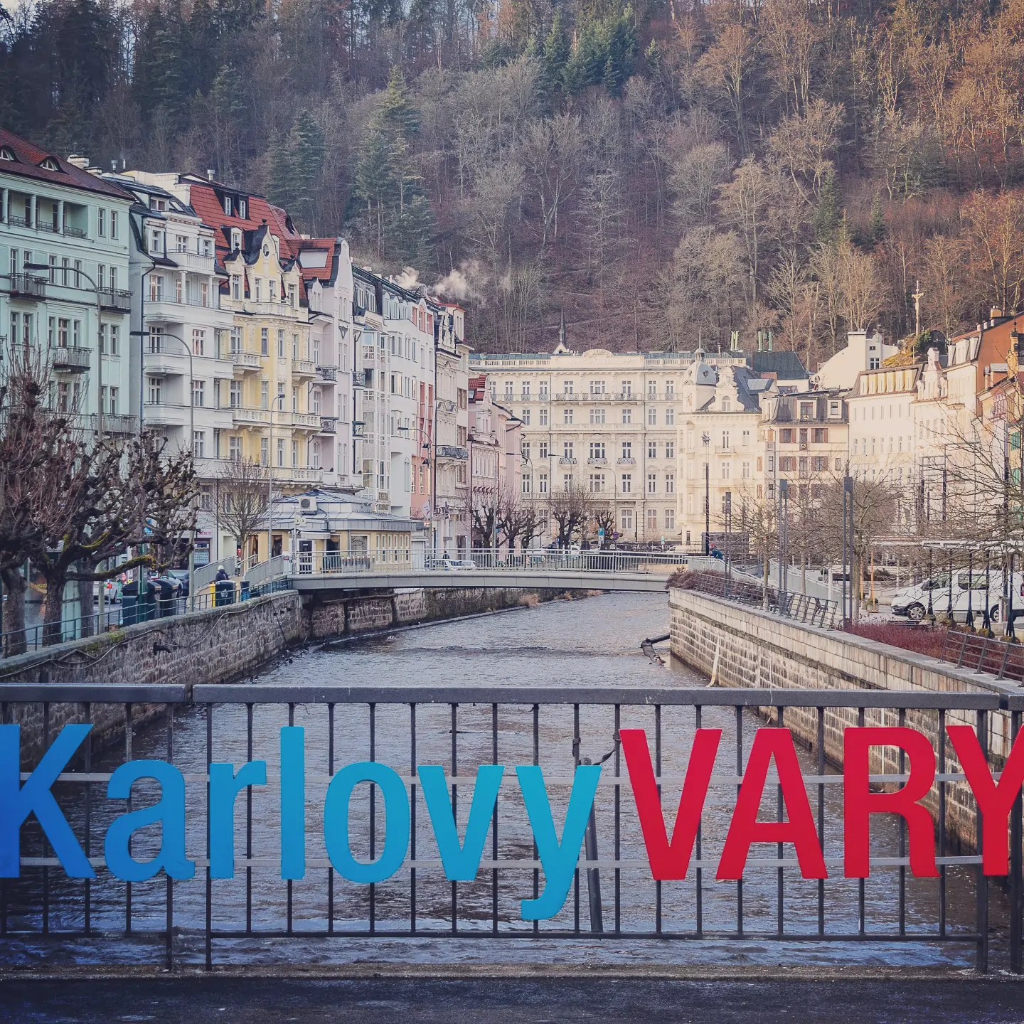 A colorful “Karlovy VARY” sign is mounted on a bridge railing above a river lined with pastel buildings and wooded hills.