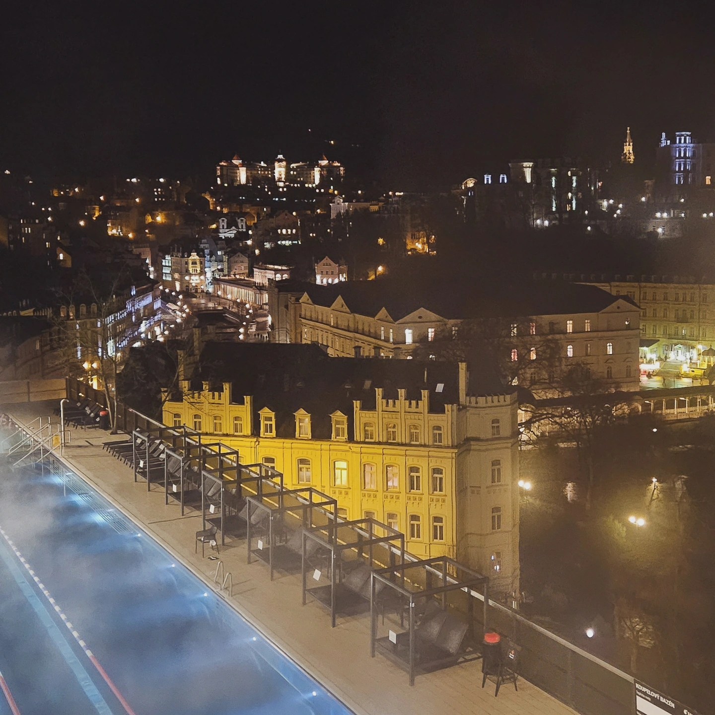A nighttime view over Karlovy Vary showing illuminated historic buildings, a rooftop terrace with a heated pool in the foreground, and softly lit streets below.