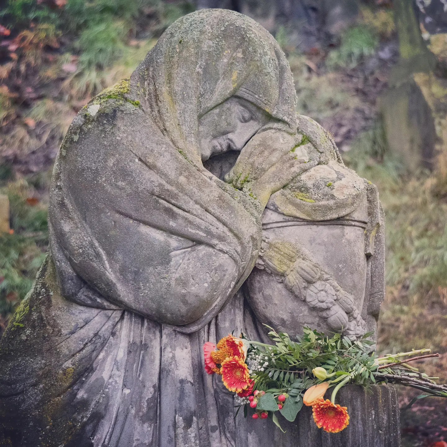A weathered stone sculpture of a cloaked figure resting its head on folded arms, with a bouquet of orange and red flowers placed at the base.