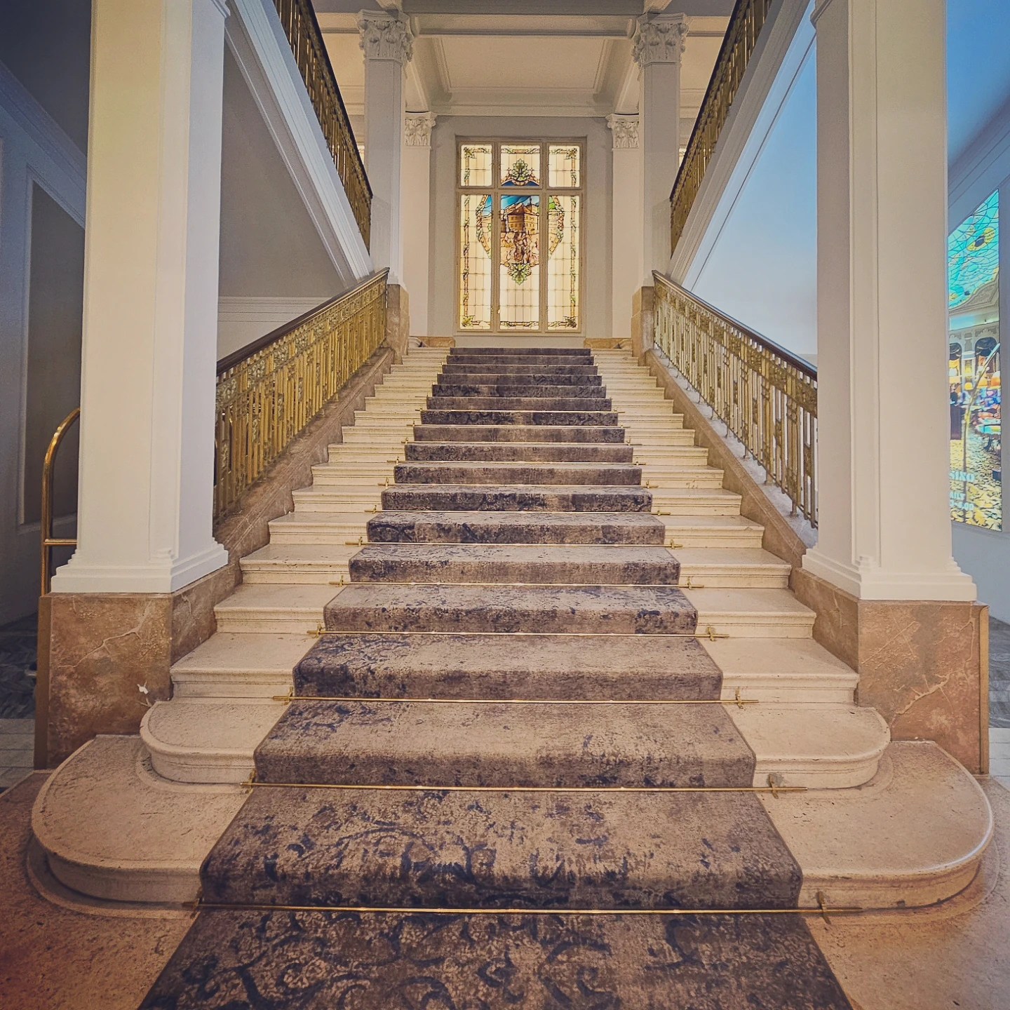 A wide marble staircase with a patterned runner and gilded railings leading up to a stained glass window inside Grandhotel Pupp.