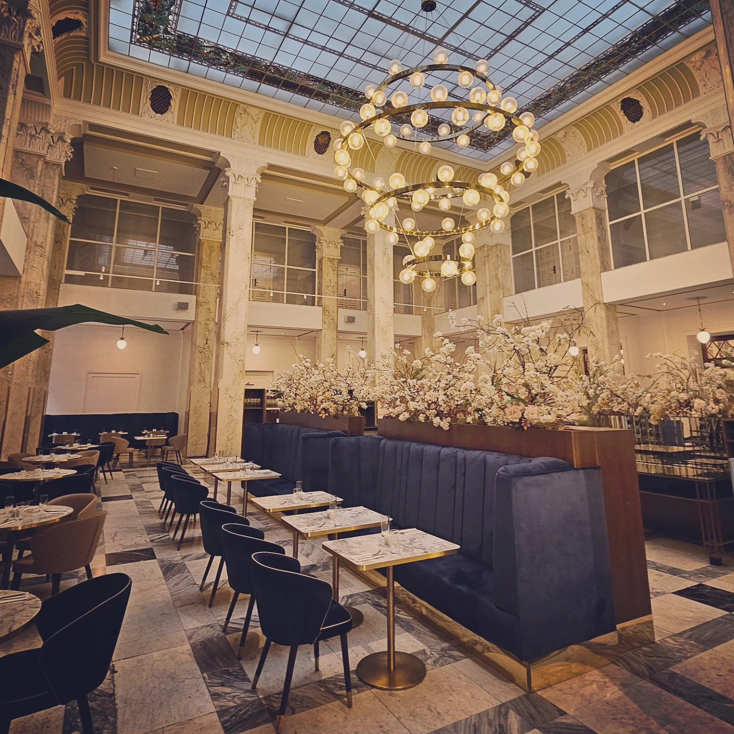An elegant restaurant hall inside Grandhotel Pupp with marble columns, a glass ceiling, blue velvet seating, and a large circular chandelier.