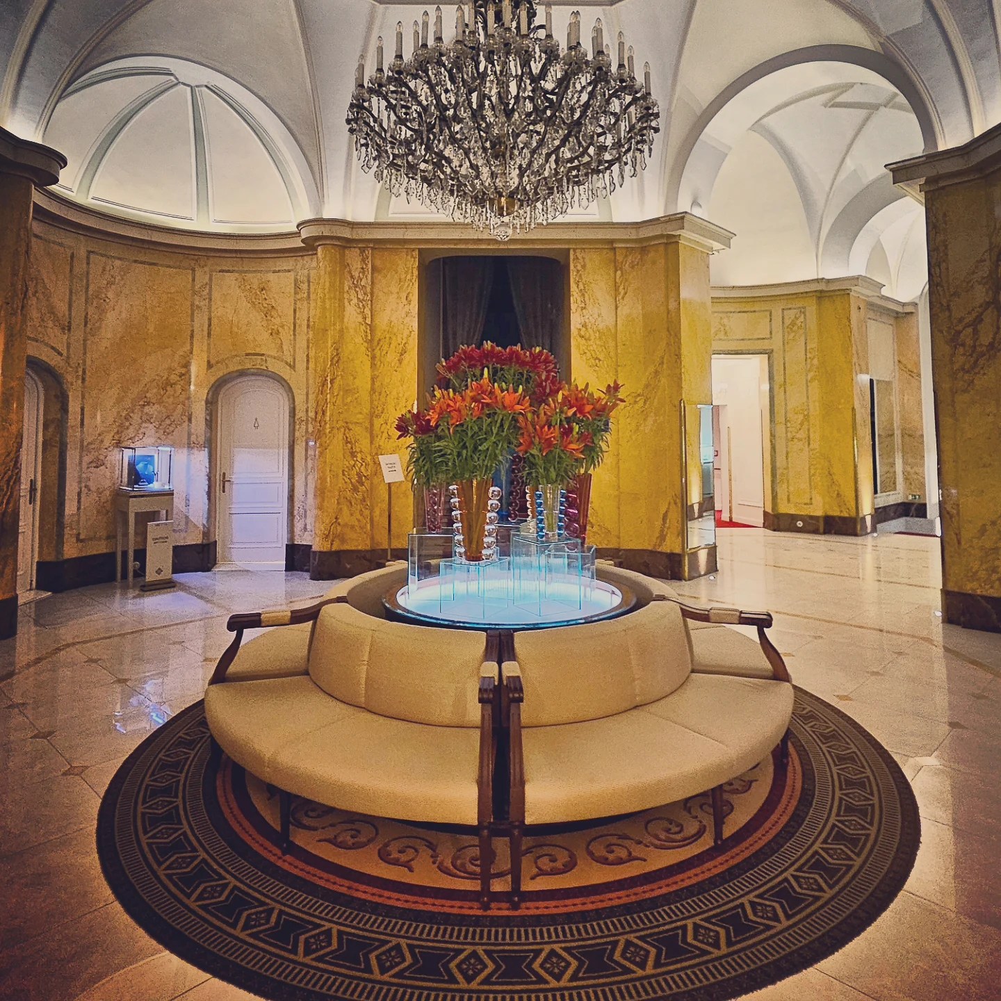 A circular seating arrangement around a glass display with flowers beneath a crystal chandelier in the lobby of Grandhotel Pupp.
