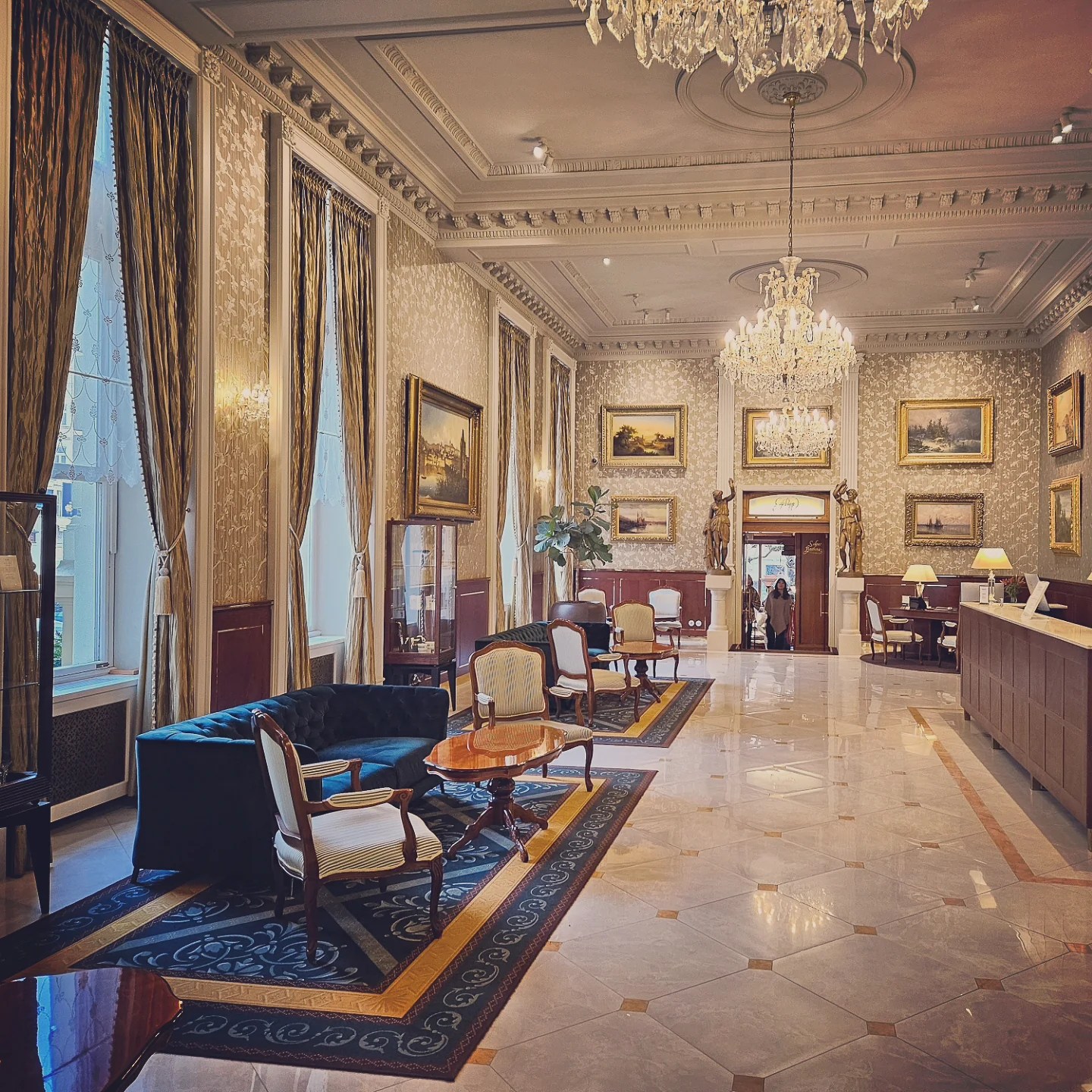 A refined lounge inside Grandhotel Pupp with chandeliers, framed paintings, tall windows with drapes, and classic upholstered seating.