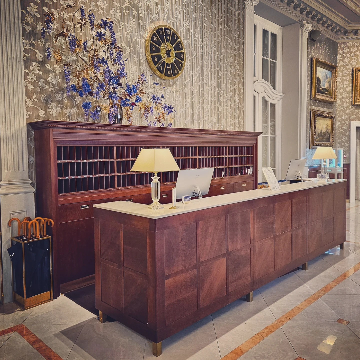 The wooden reception desk at Grandhotel Pupp with a wall of mail slots, a gold clock above, and a vase of blue flowers.