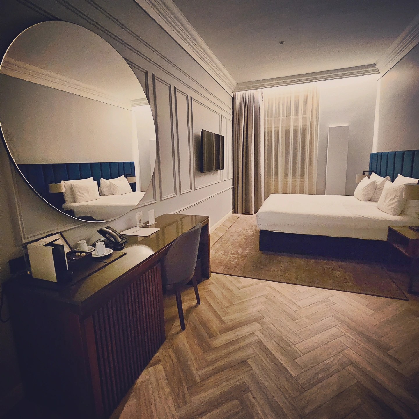 A modern guest room at Grandhotel Pupp with a large bed, blue upholstered headboard, round wall mirror, desk, and parquet wood flooring.