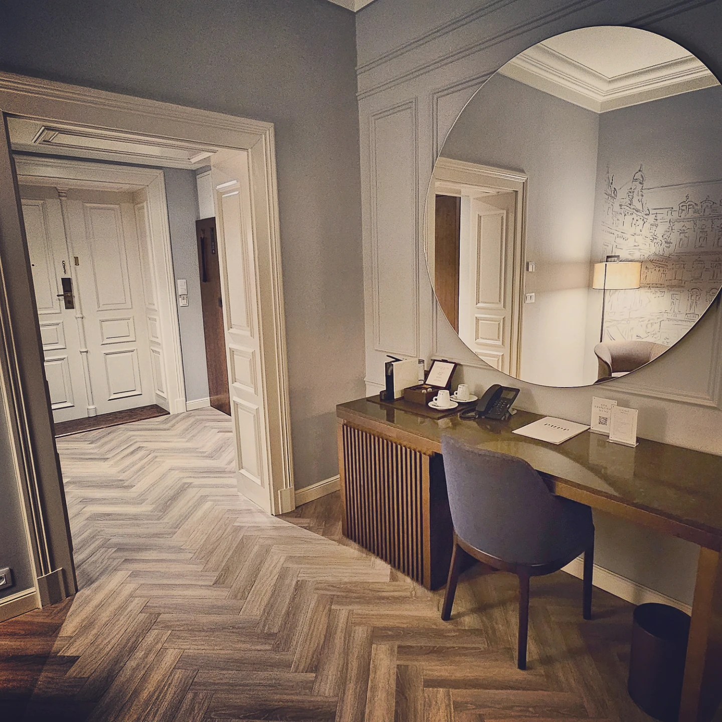 A hotel room entry area at Grandhotel Pupp with a wooden desk, round mirror, patterned herringbone flooring, and white paneled doors.