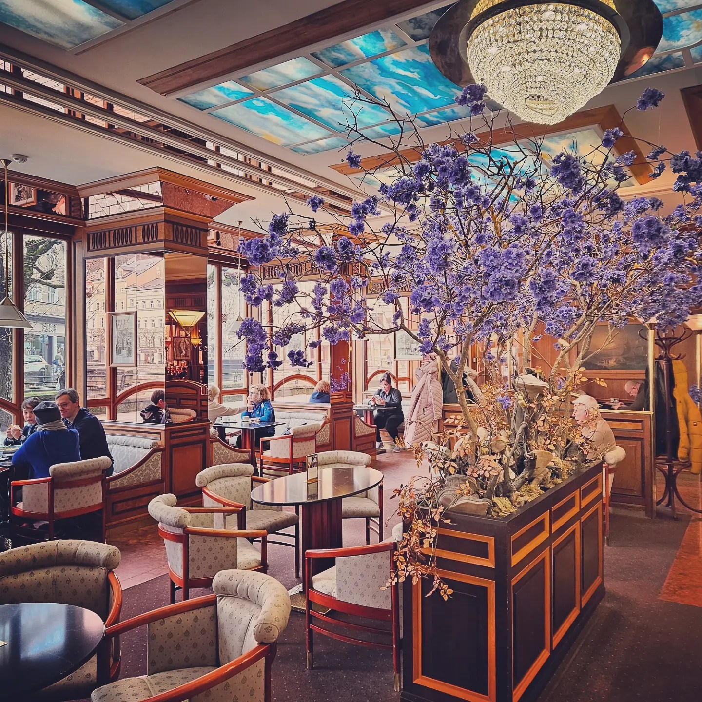 The Café Pupp interior at Grandhotel Pupp with small round tables, upholstered chairs, a large floral arrangement of purple blossoms, and a decorative glass ceiling.