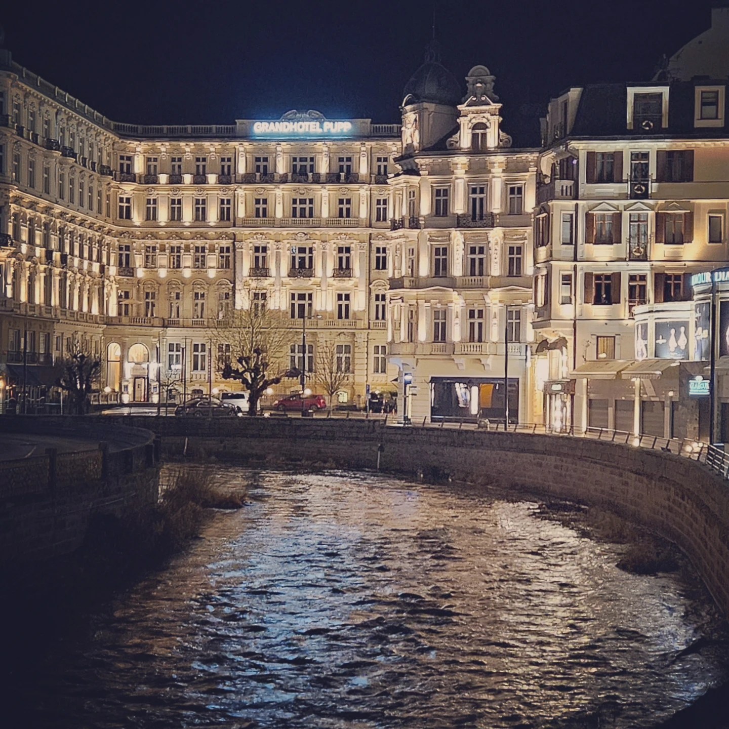 Grandhotel Pupp illuminated at night along the Teplá River in Karlovy Vary, with its historic façade glowing against the dark sky.