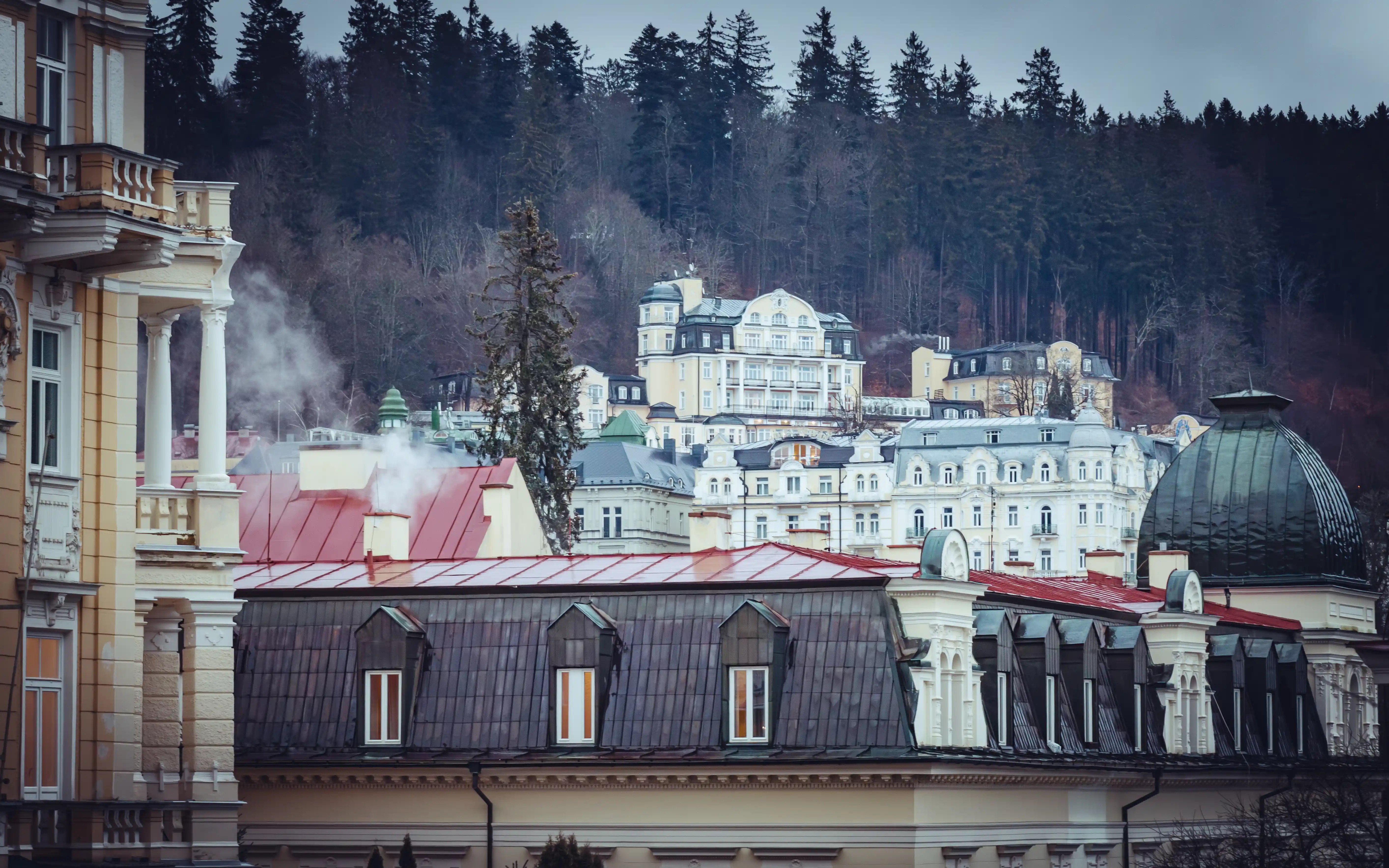 A layered view of historic pastel spa buildings with red and green rooftops set against a forested hillside in Mariánské Lázně.