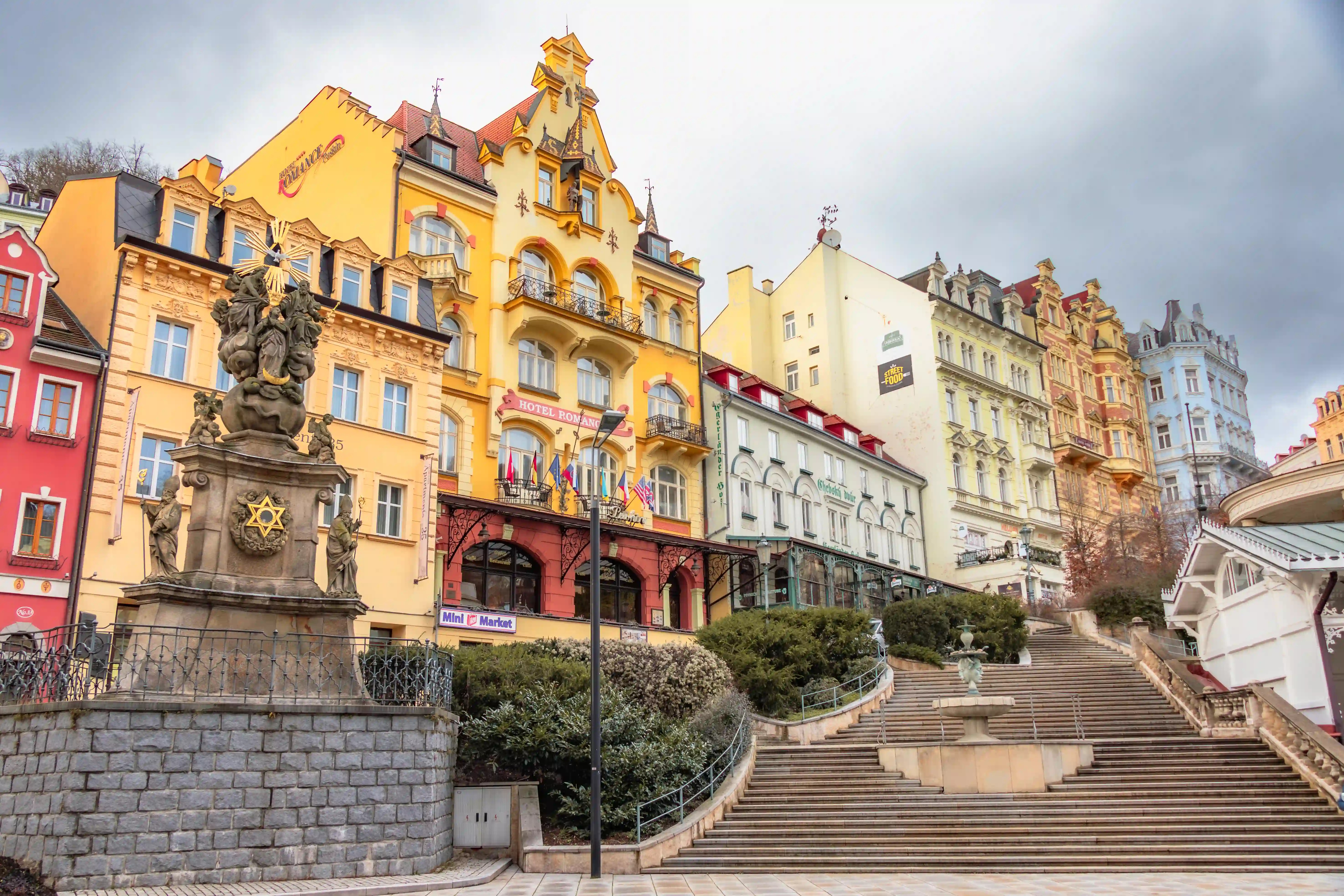 A wide staircase in Karlovy Vary leading up to colorful historic buildings, with a large Baroque statue standing on a stone pedestal to the left.