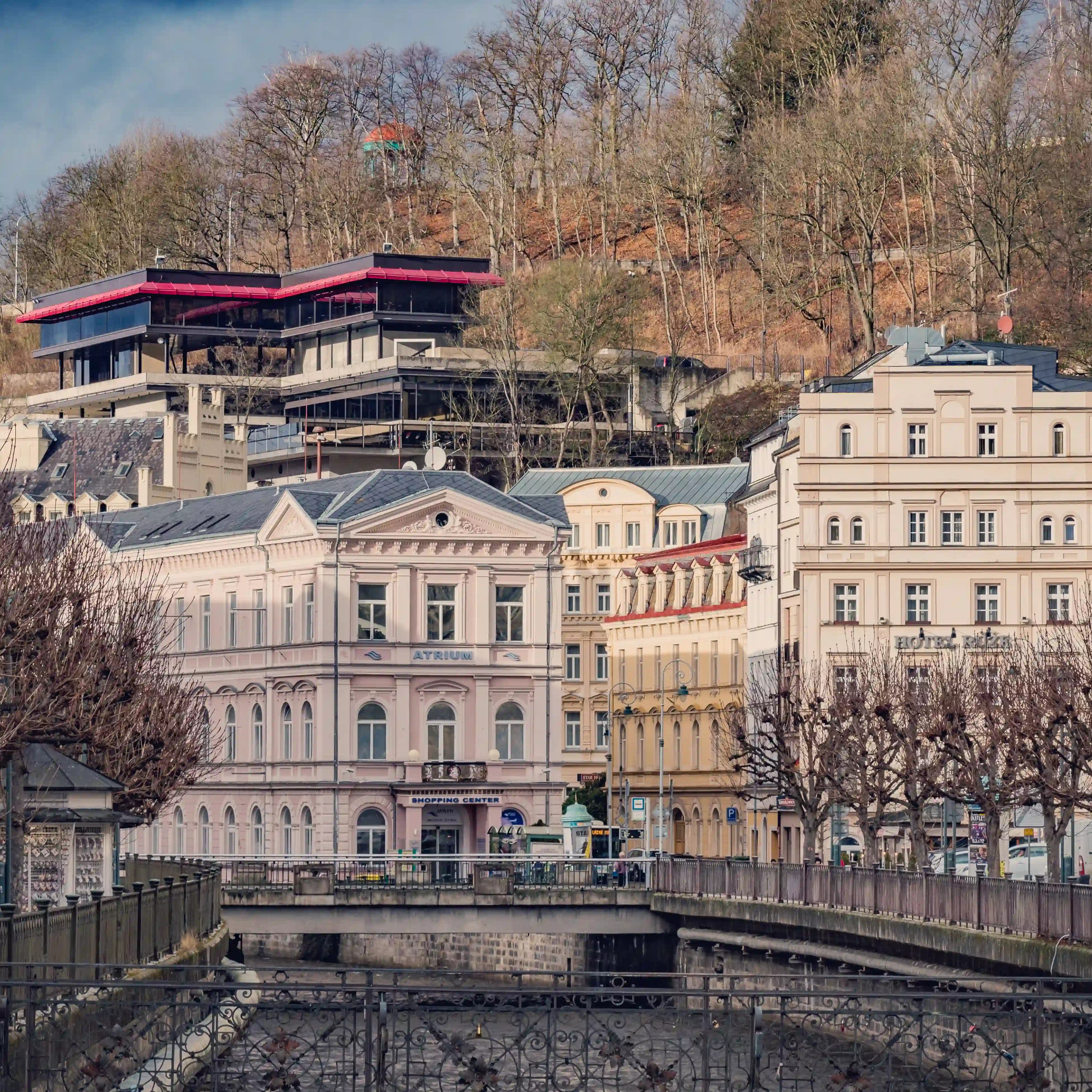 The Atrium Shopping Center building and the Saunia Thermal Resort in Karlovy Vary viewed across a river canal with ornate railings and leafless trees lining the banks.