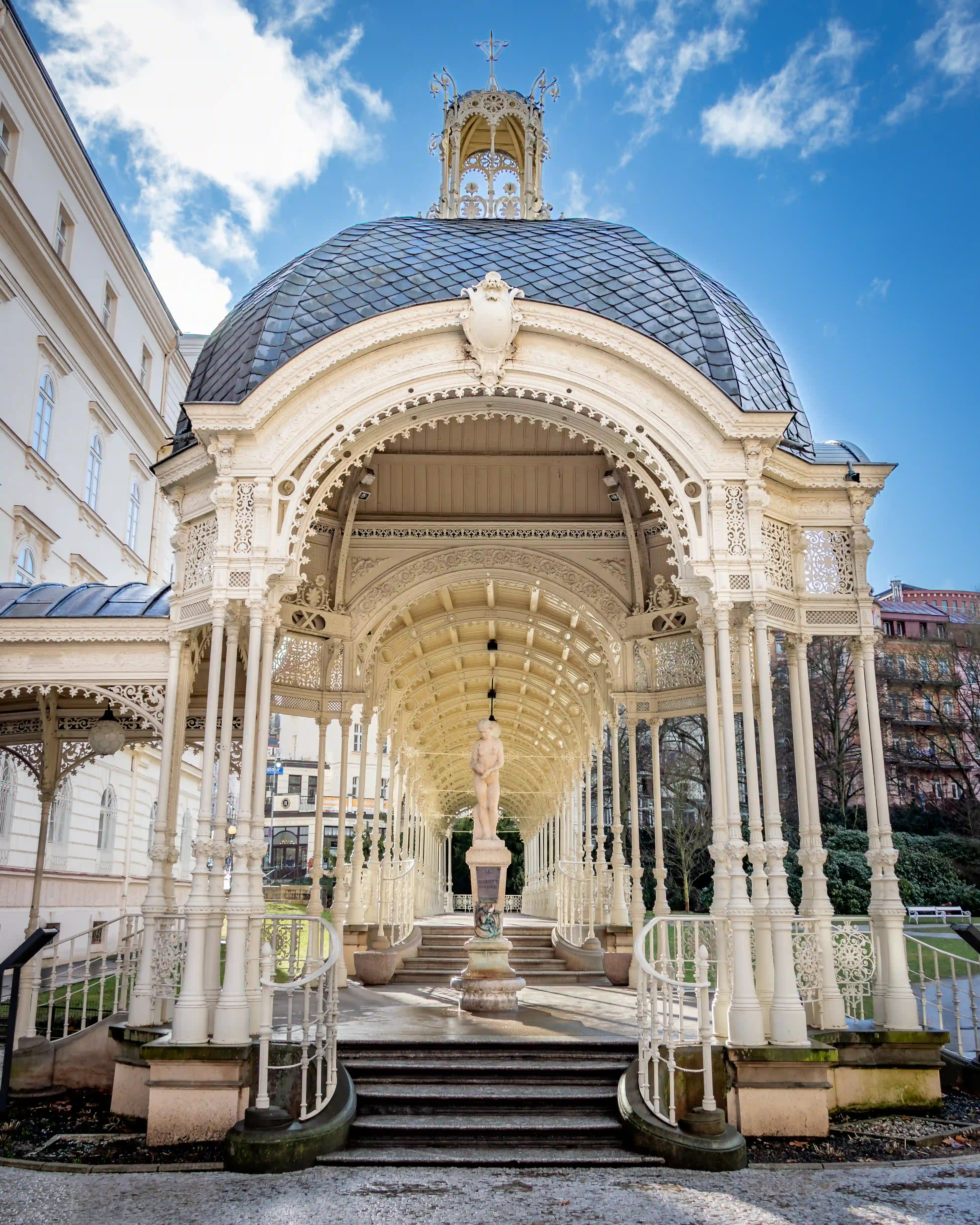 A stone statue stands centered beneath an elaborately decorated cream colonnade with curved staircases.