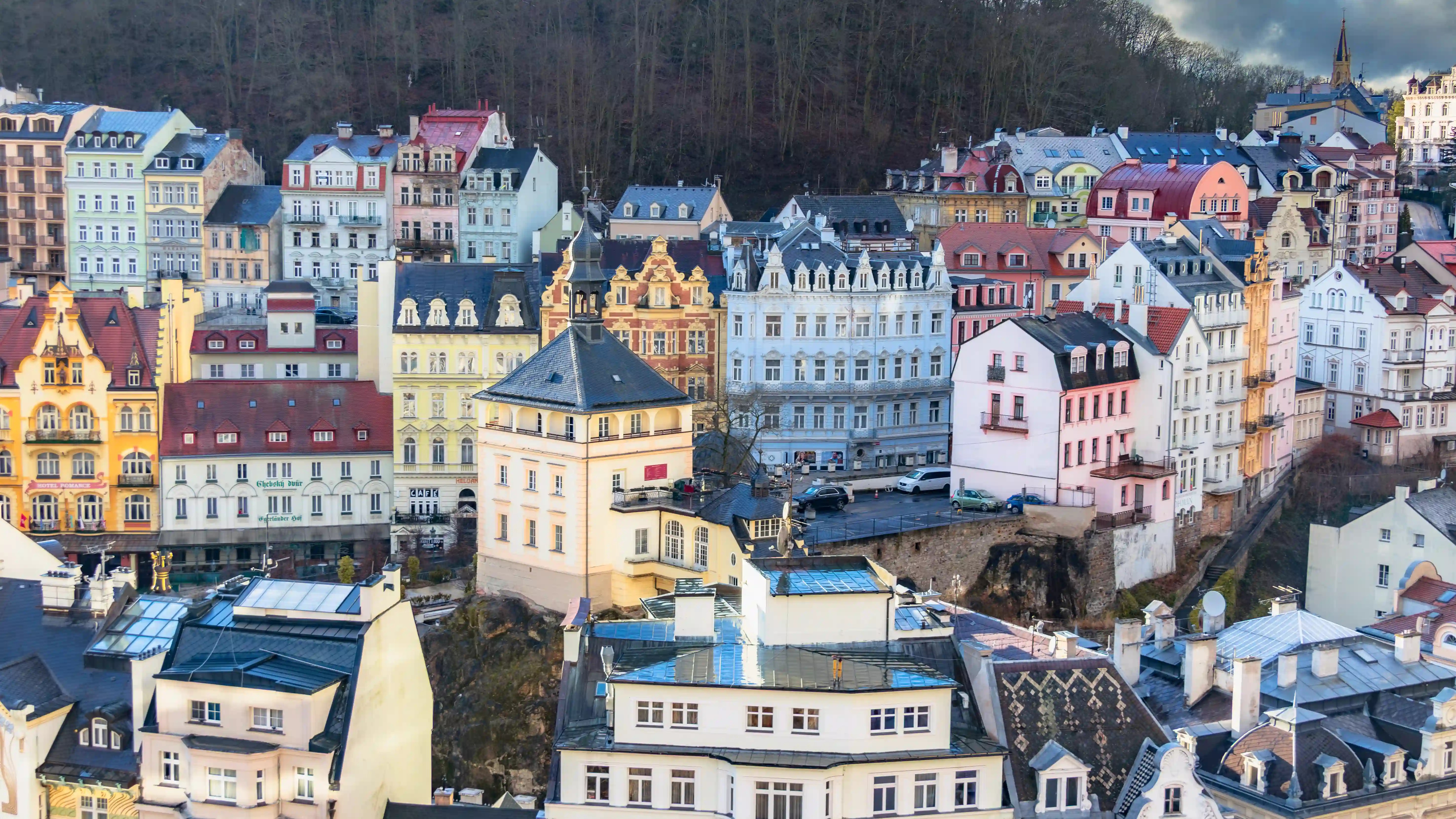 Colorful historic buildings clustered on a hillside above Karlovy Vary’s old town, with ornate facades and red rooftops beneath a wooded slope.
