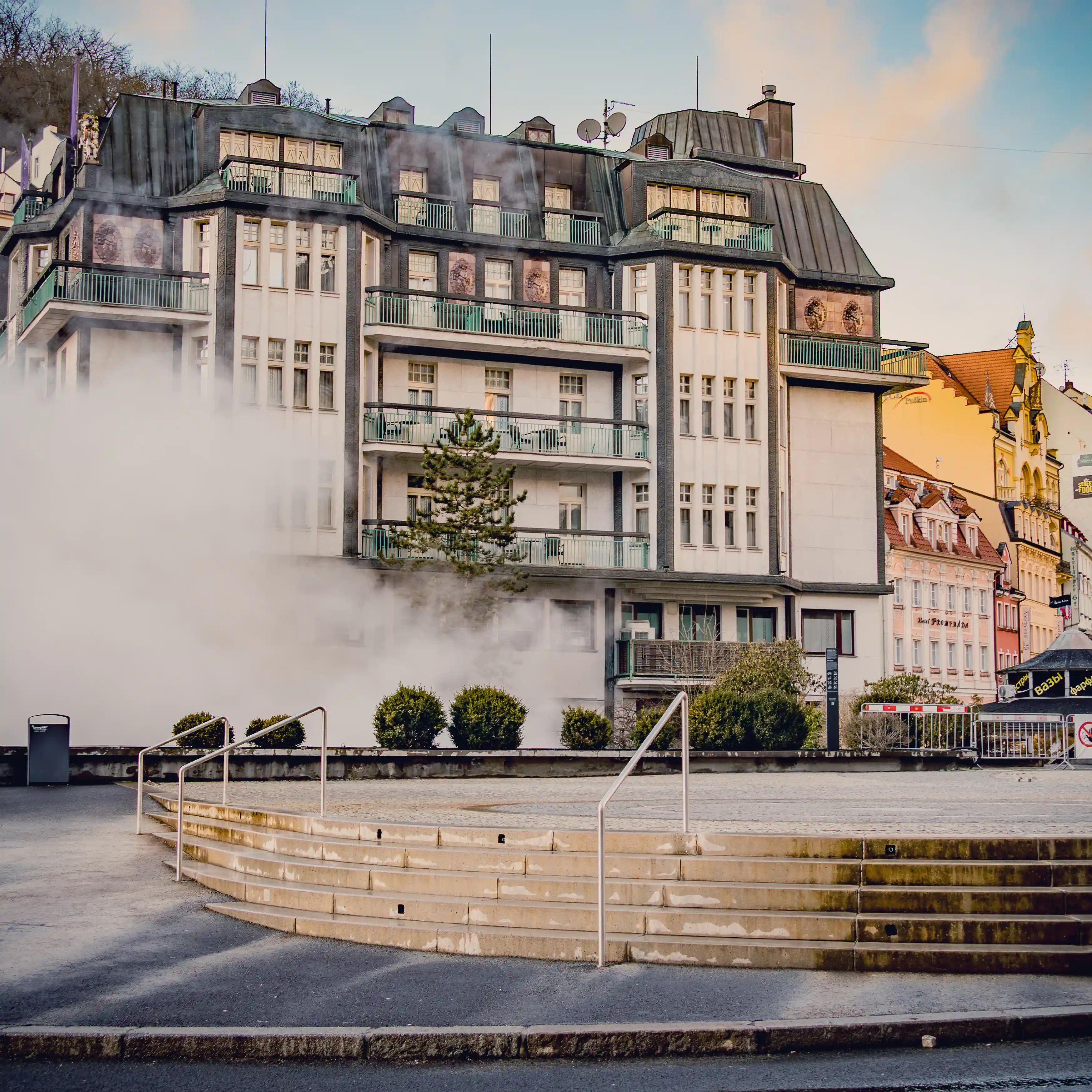 Thick steam rises from a hot spring in Karlovy Vary, partially obscuring the buildings behind it.