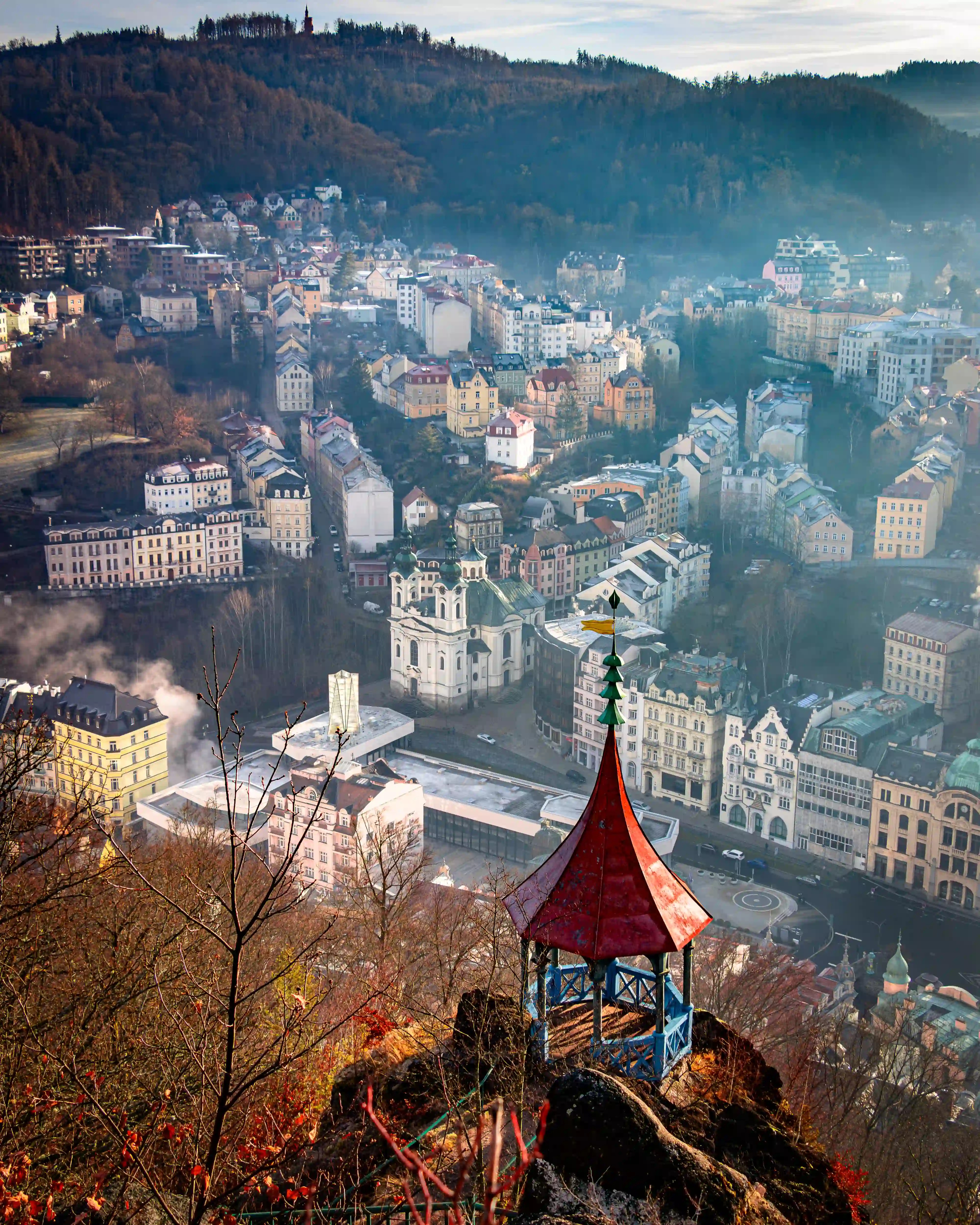 A red-roofed gazebo perched on a rocky outcrop overlooking Karlovy Vary’s pastel buildings and mist-filled valley.