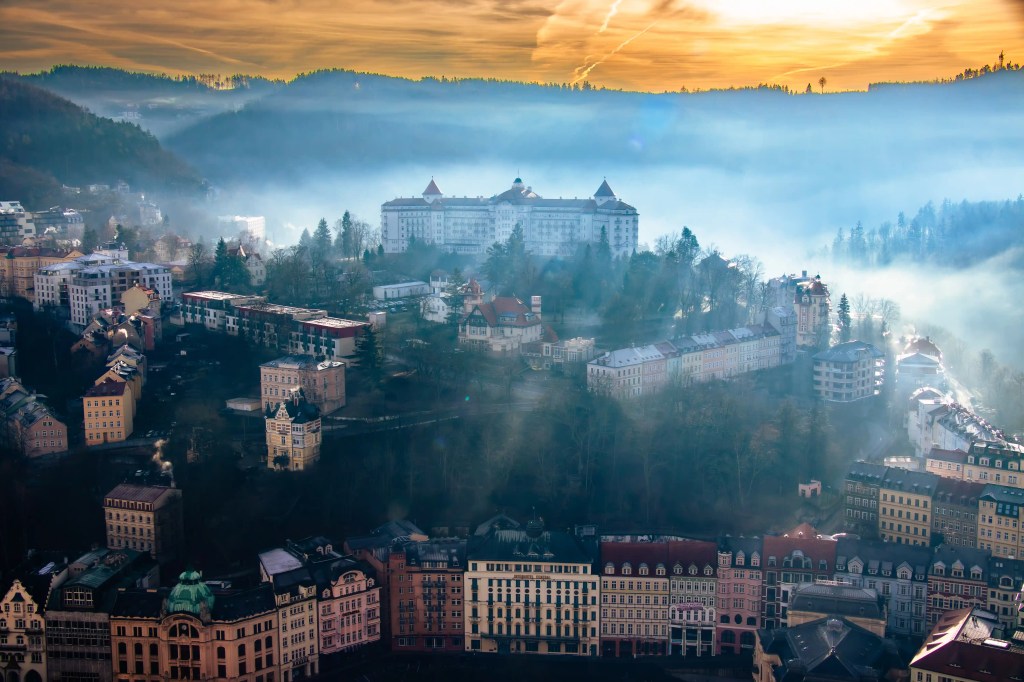 A foggy sunrise view of Karlovy Vary with rows of ornate buildings along the river and a large hilltop hotel in the background.