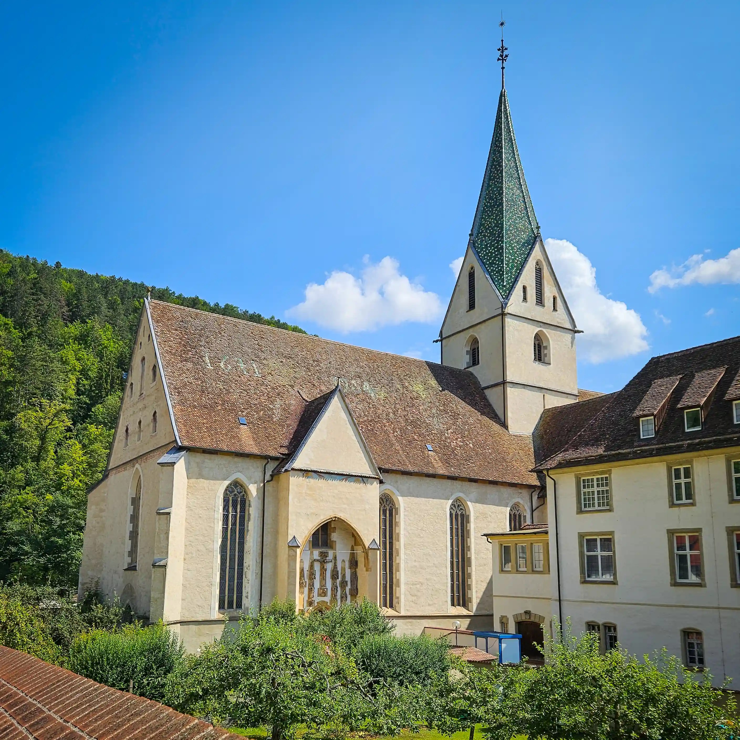 The cream-colored church of Kloster Blaubeuren with a tall green spire rises beside orchard trees and forested hills.