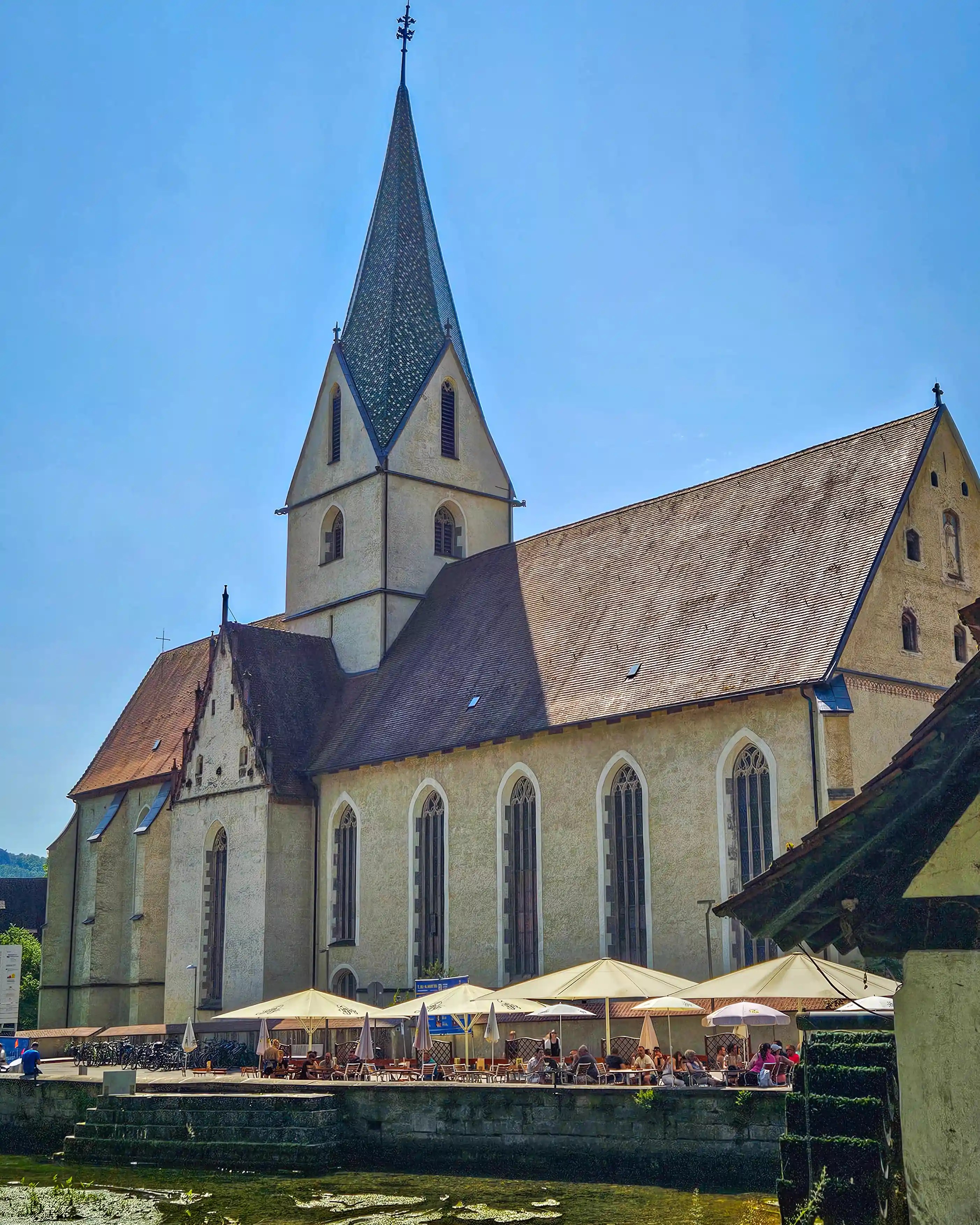 Kloster Blaubeuren’s church stands beside the Blautopf, with café tables and umbrellas along the water’s edge.