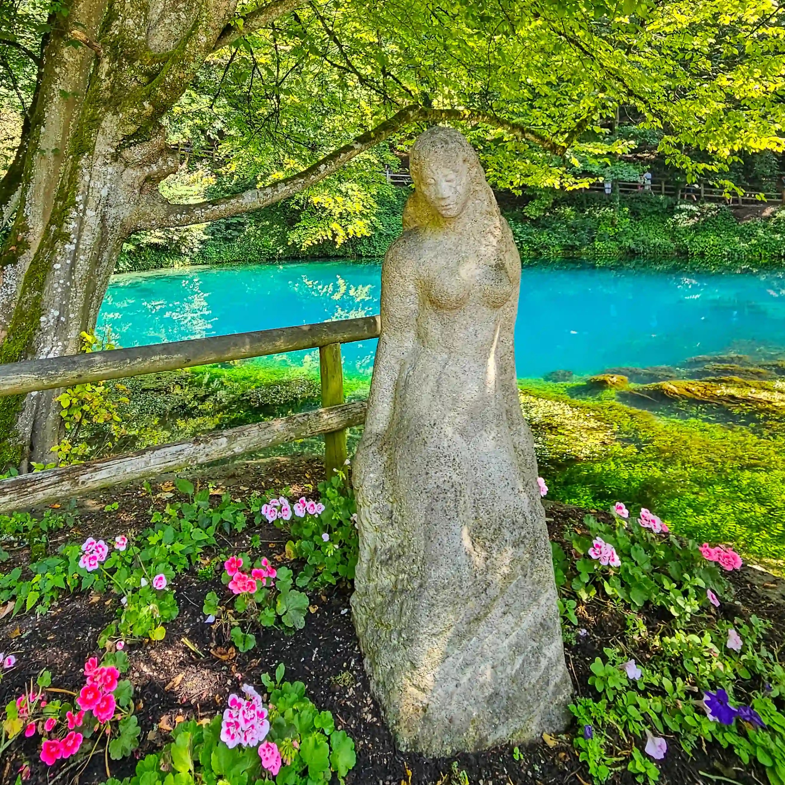 A stone statue of a woman stands beside the turquoise Blautopf, surrounded by pink flowers and shaded by trees.