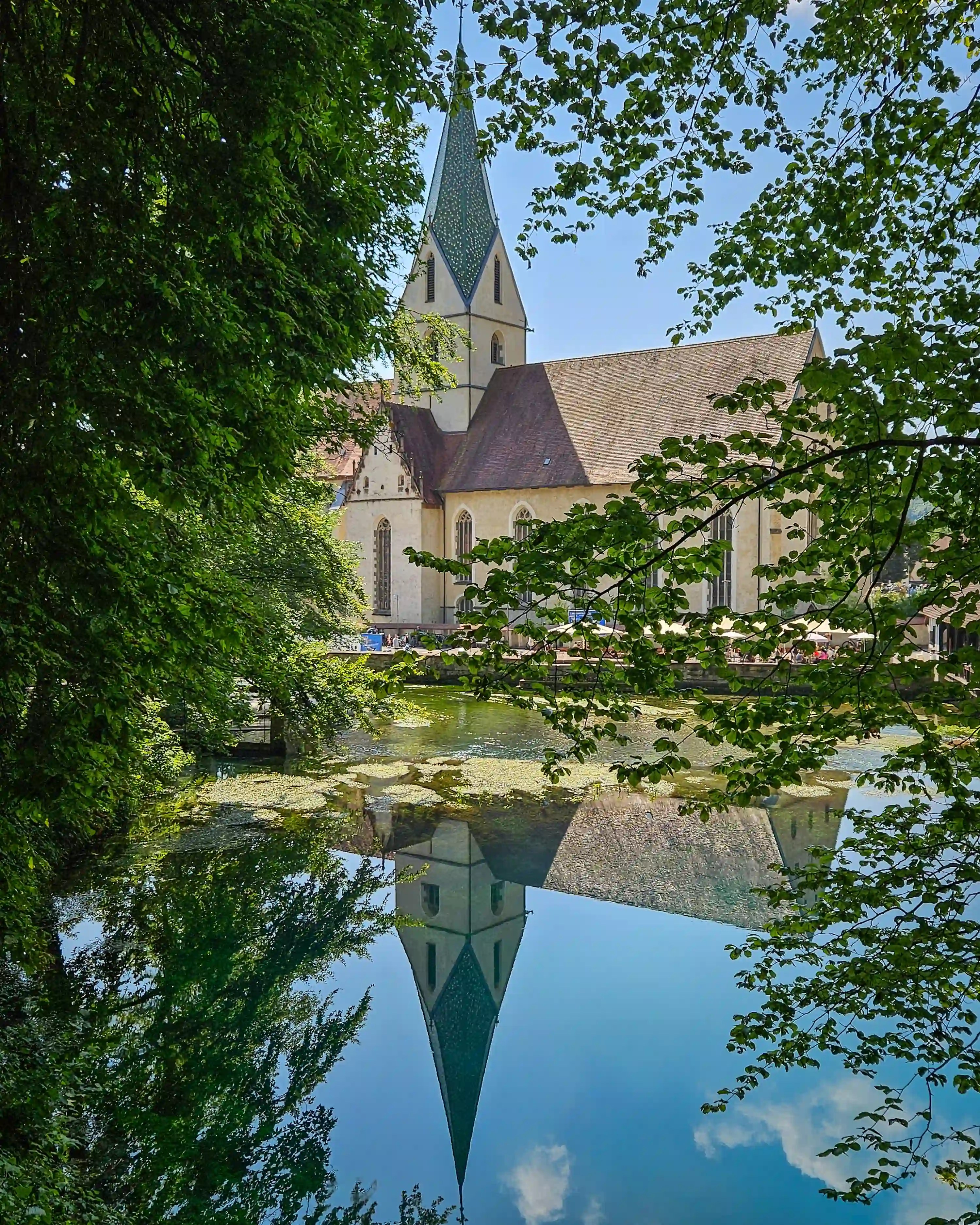 Blaubeuren Abbey Church rises above the water, its tall spire reflected clearly in the calm surface of the Blautopf.