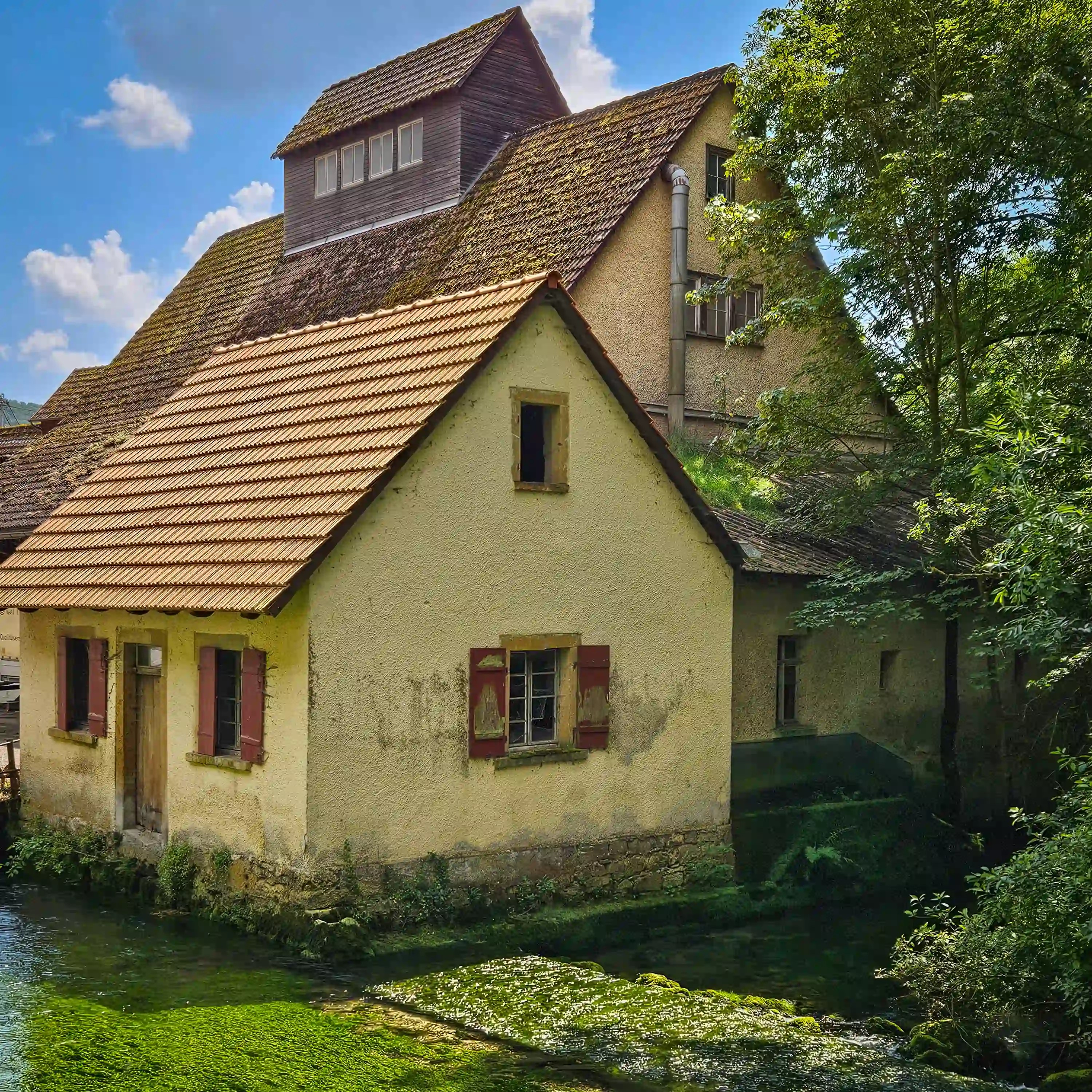 A small yellow house with red shutters stands directly over clear green water beside the Blautopf.