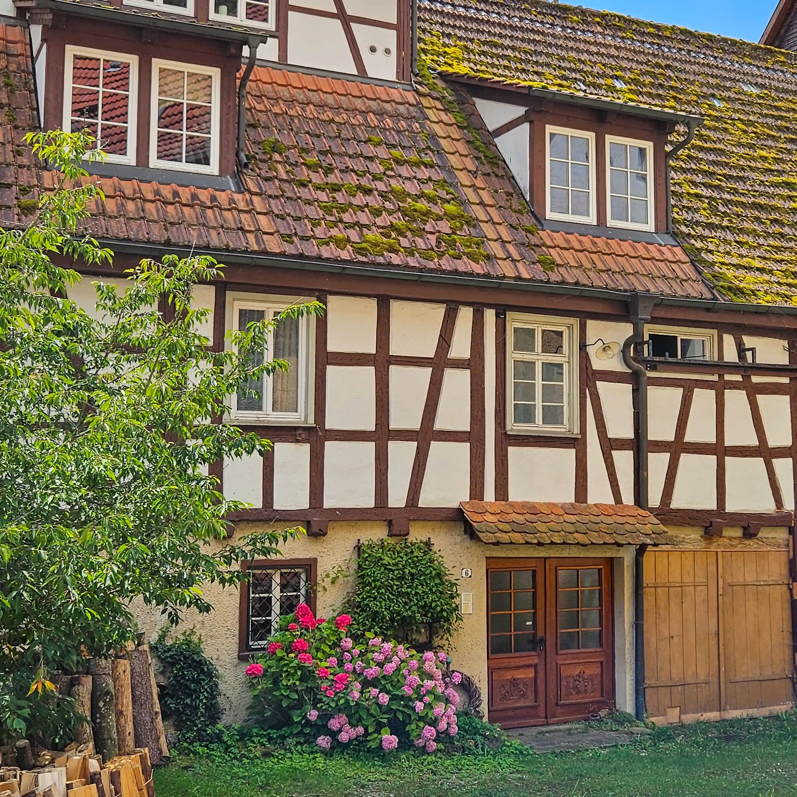 A traditional half-timbered house with a moss-covered tiled roof and pink hydrangeas grows beside a wooden doorway.