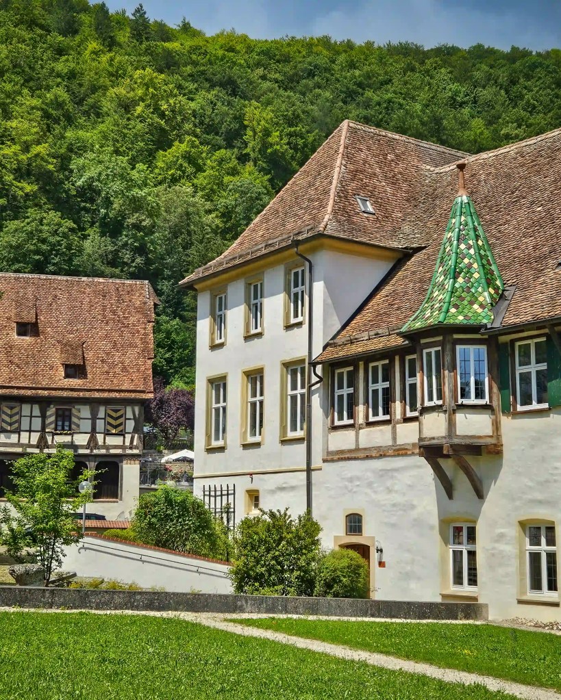 A white monastery building with a small green tiled turret and timber-framed upper story stands beside a grassy yard.