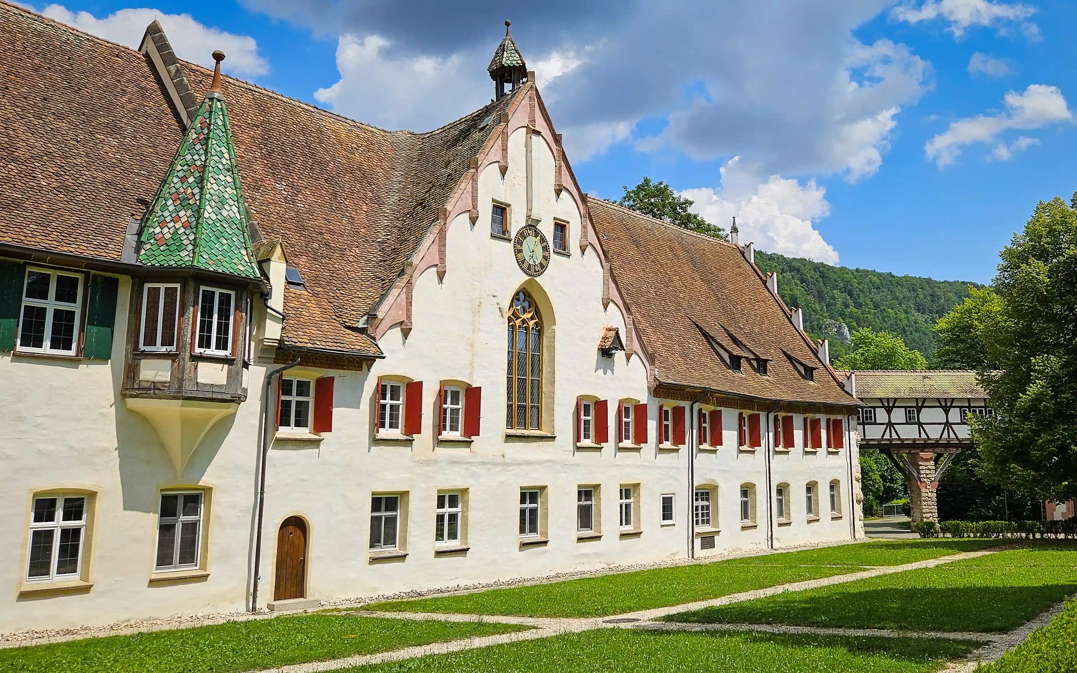 The main façade of Kloster Blaubeuren with red shutters, a clock, and Gothic windows stretches along a grassy lawn.