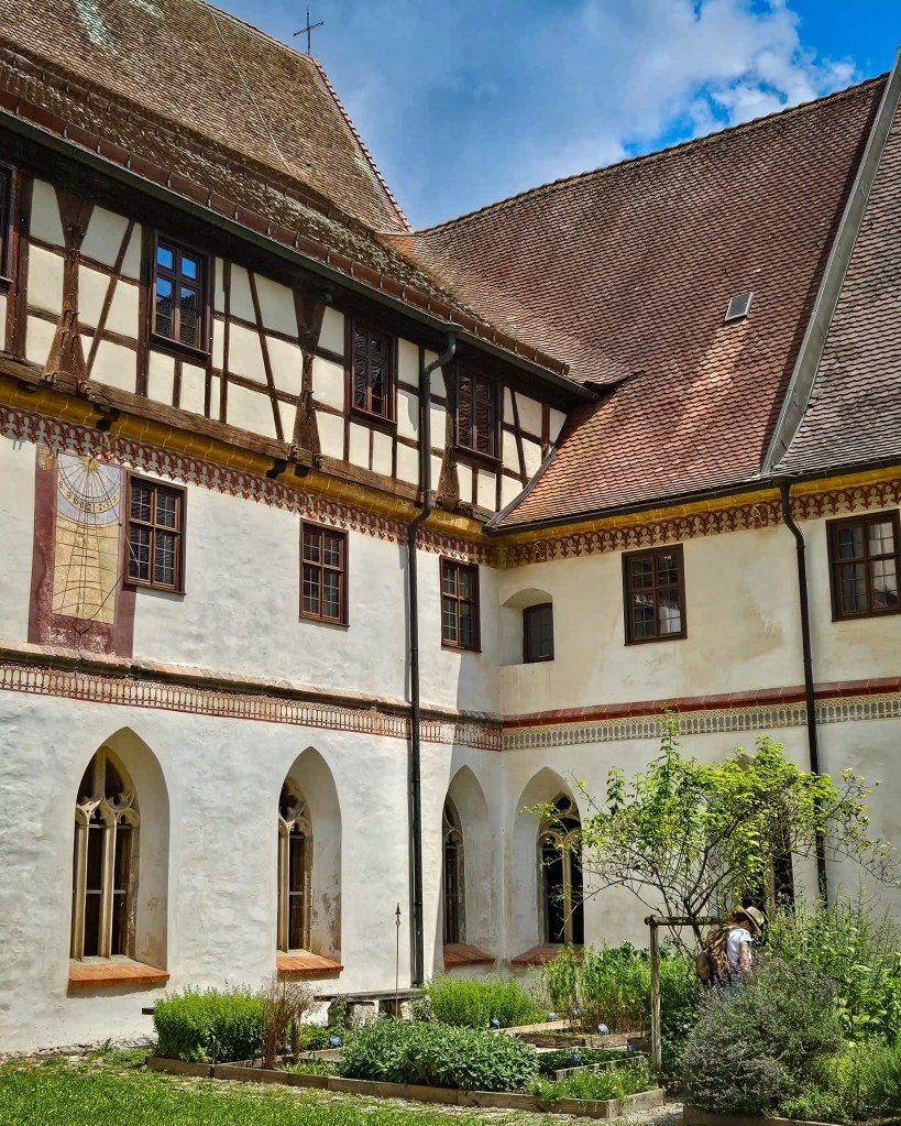The courtyard of Kloster Blaubeuren shows Gothic arched windows, a timber-framed upper level, and a painted sundial above the herb garden.