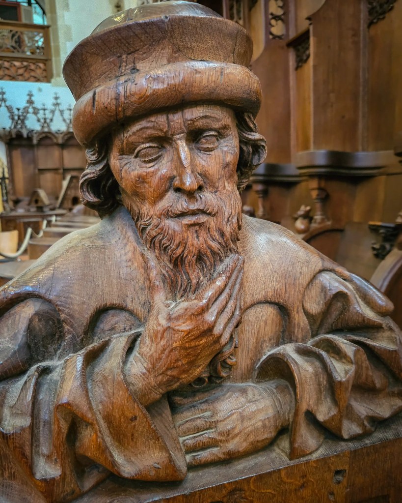 A close-up of a carved wooden figure with a beard and hat, part of the choir stalls inside Kloster Blaubeuren.