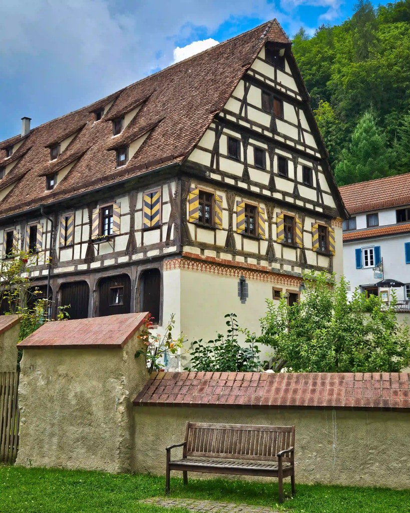 A large half-timbered building near Kloster Blaubeuren stands behind a low wall with a wooden bench in the foreground.