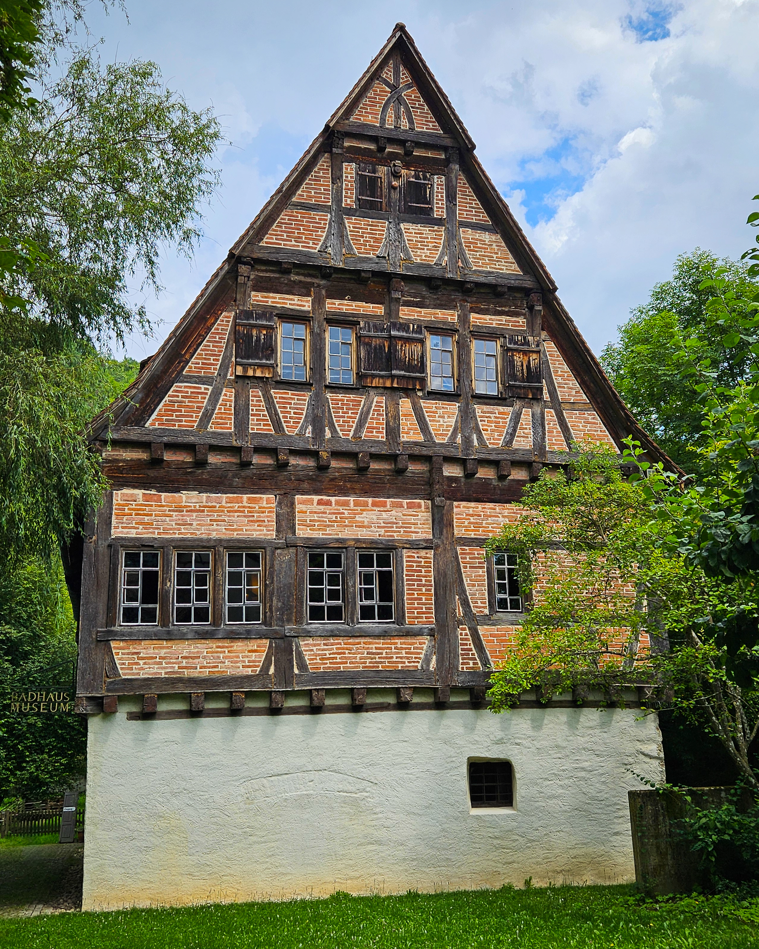 A tall half-timbered brick house with dark wooden beams and shuttered windows stands beside trees near the Blautopf in Blaubeuren.