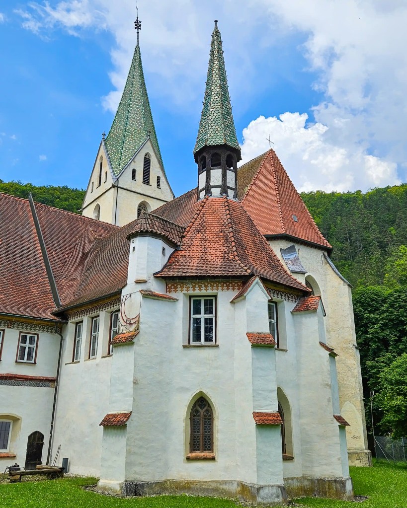 The exterior of Kloster Blaubeuren shows white plaster walls, red-tiled roofs, and two green spires rising against the hills.
