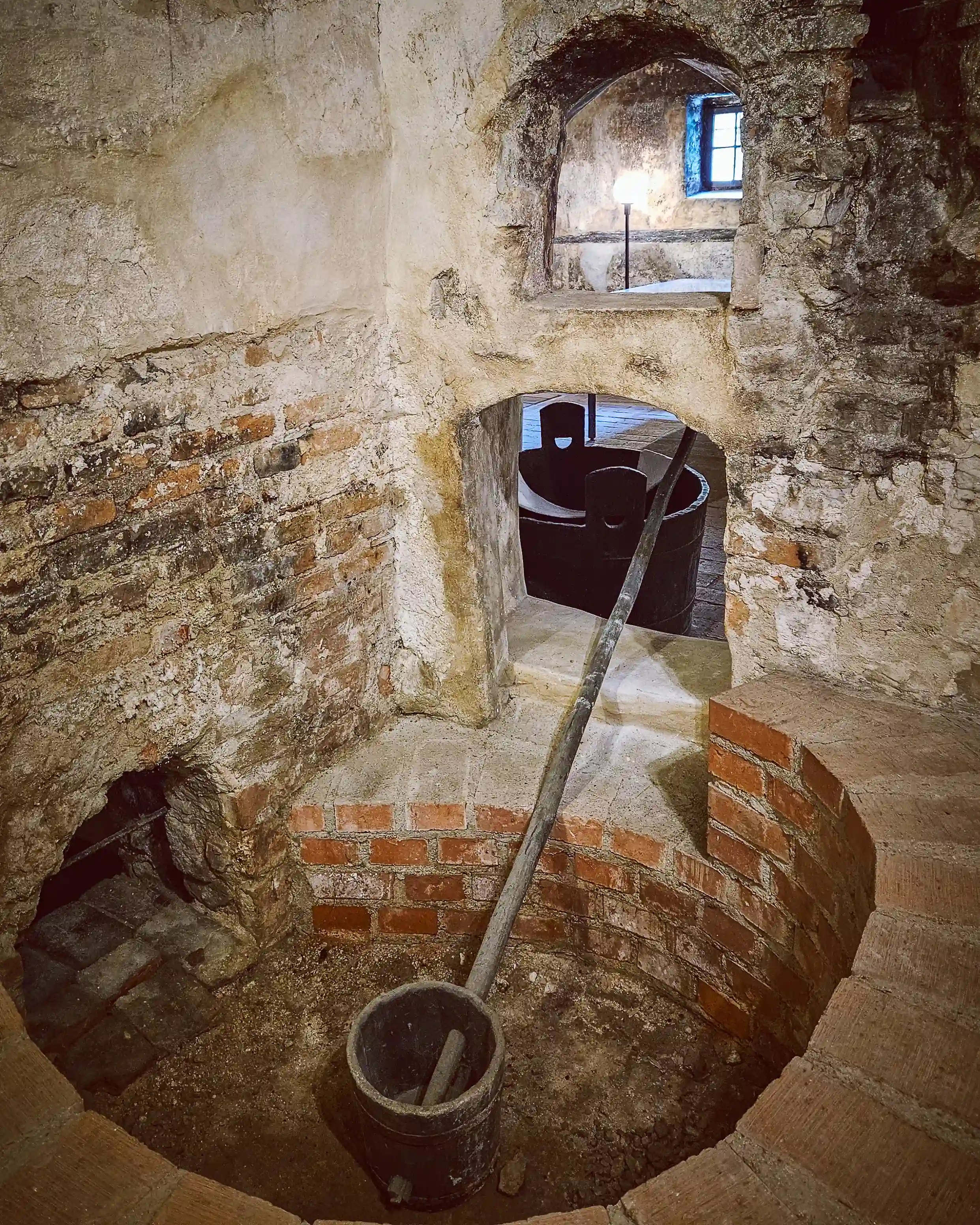 A stone well with a wooden bucket and long handle sits inside a small arched chamber with exposed brick and plaster walls.