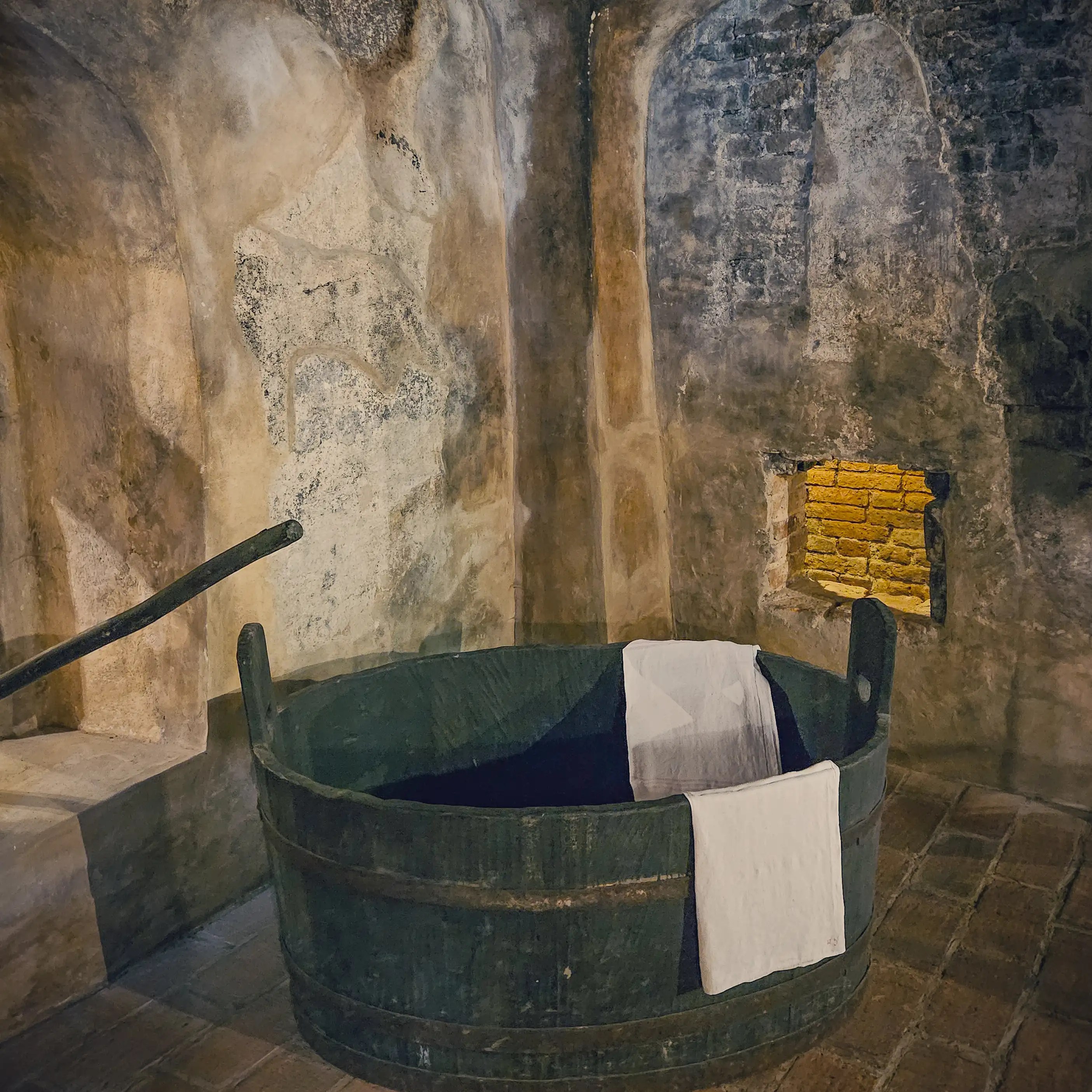 A large round wooden bathing tub with white towels draped over the side stands in a dim stone room with rough plastered walls.