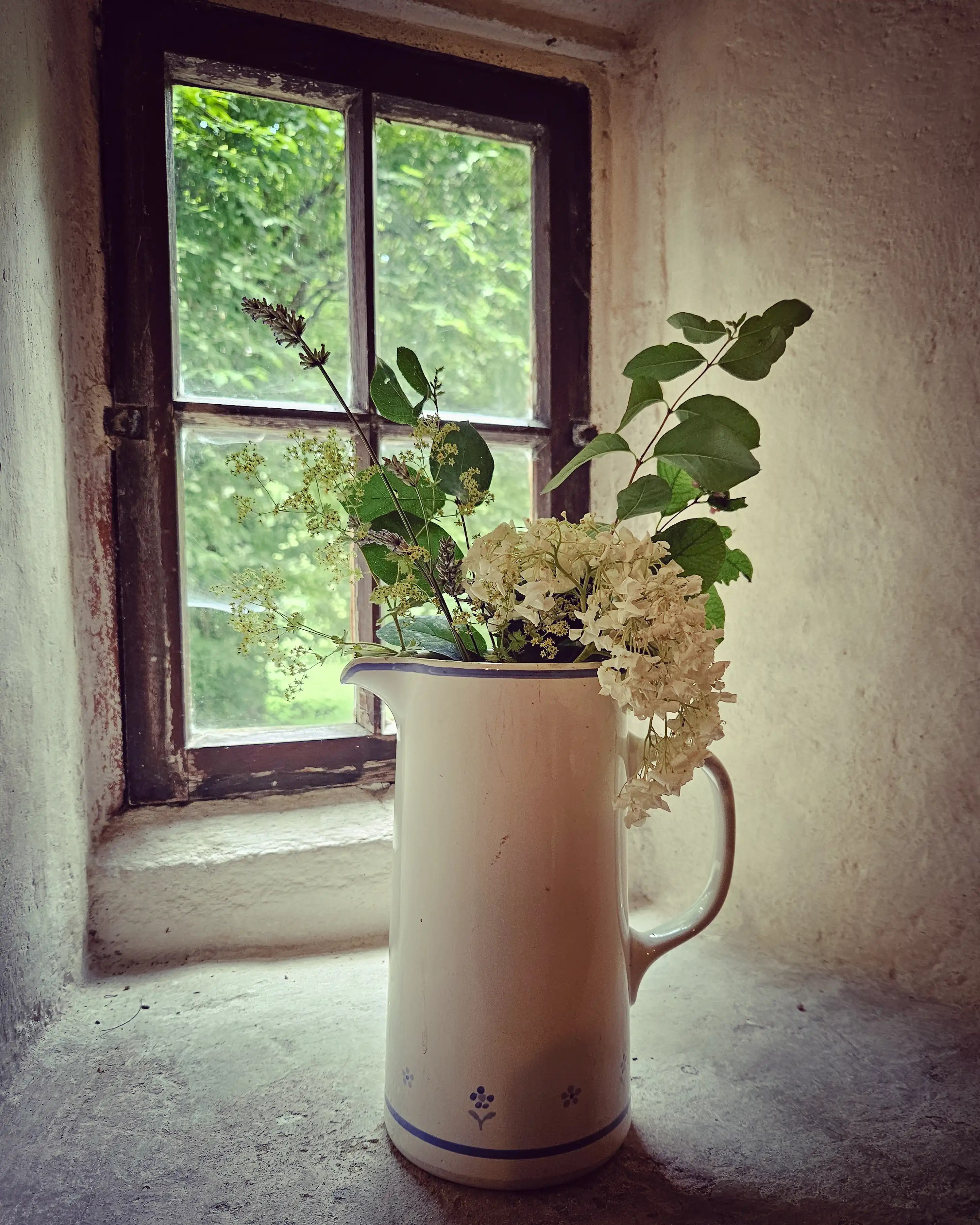 A white-colored pitcher filled with fresh flowers sits on a stone windowsill in front of a small wooden-framed window.