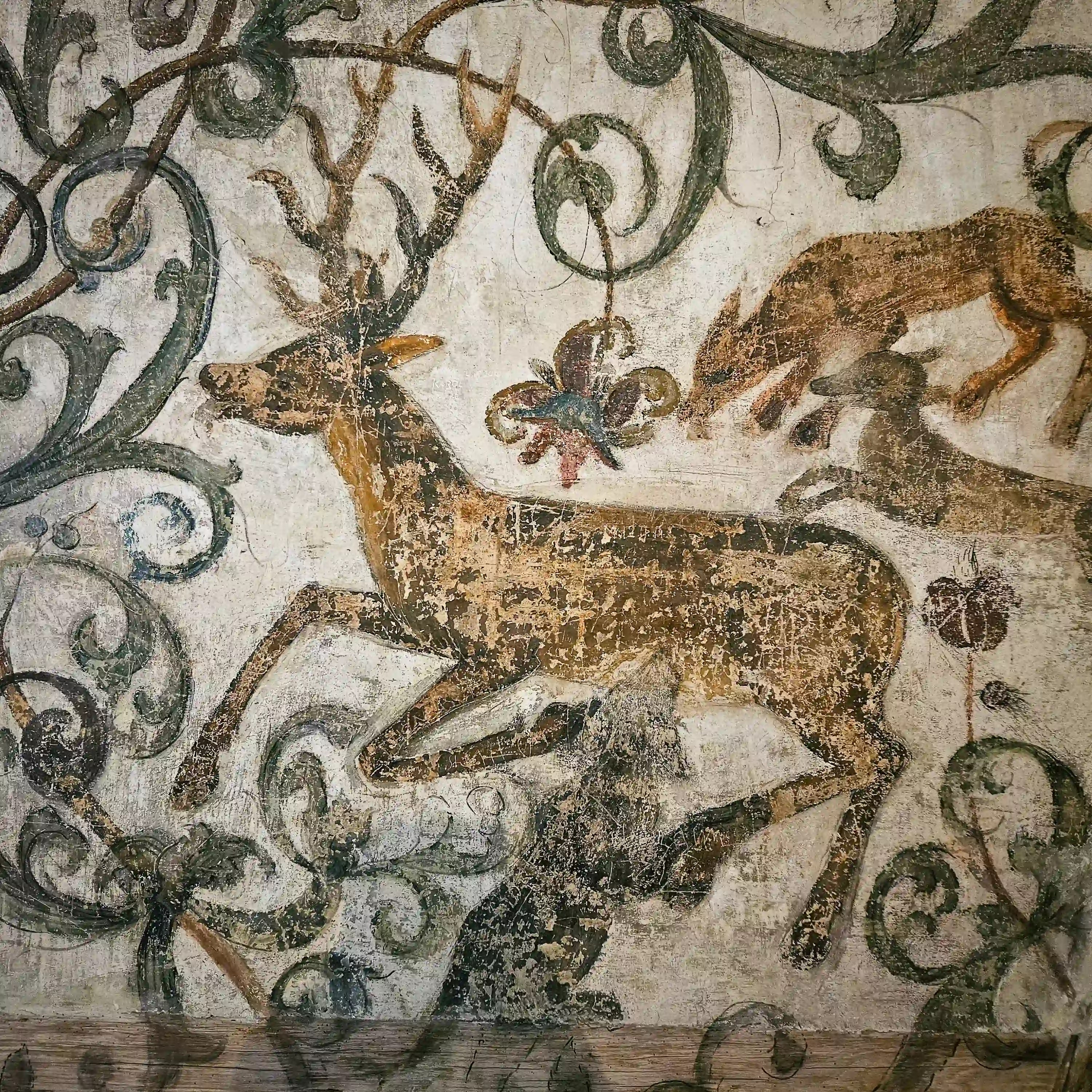 A medieval wall fresco depicts a deer with antlers surrounded by curling green vines and floral motifs.