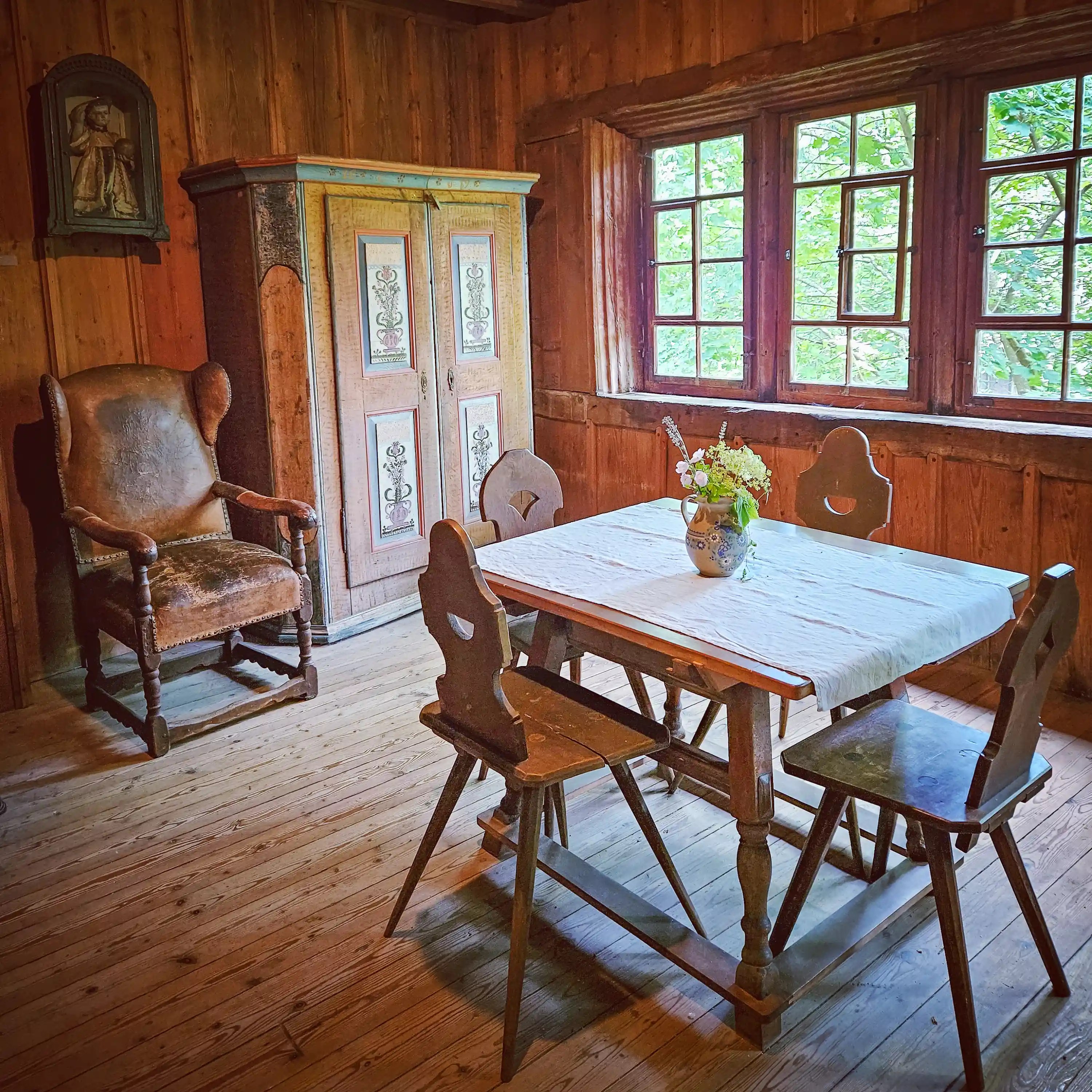 A wooden dining table with carved chairs stands in a wood-paneled room with a painted cabinet and a window overlooking greenery.