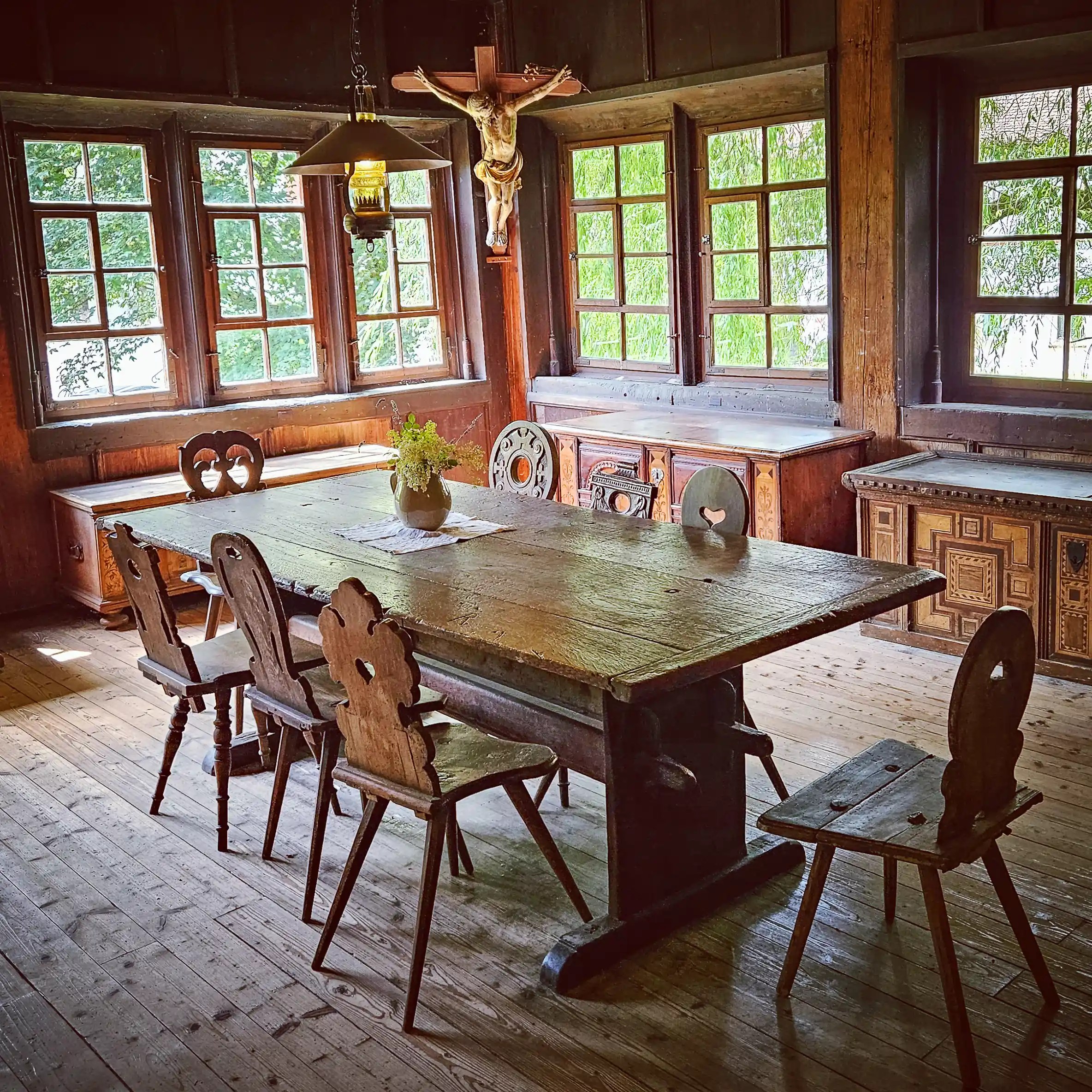 A long wooden dining table with carved chairs sits beneath a hanging lamp and crucifix in a paneled room with multiple windows.