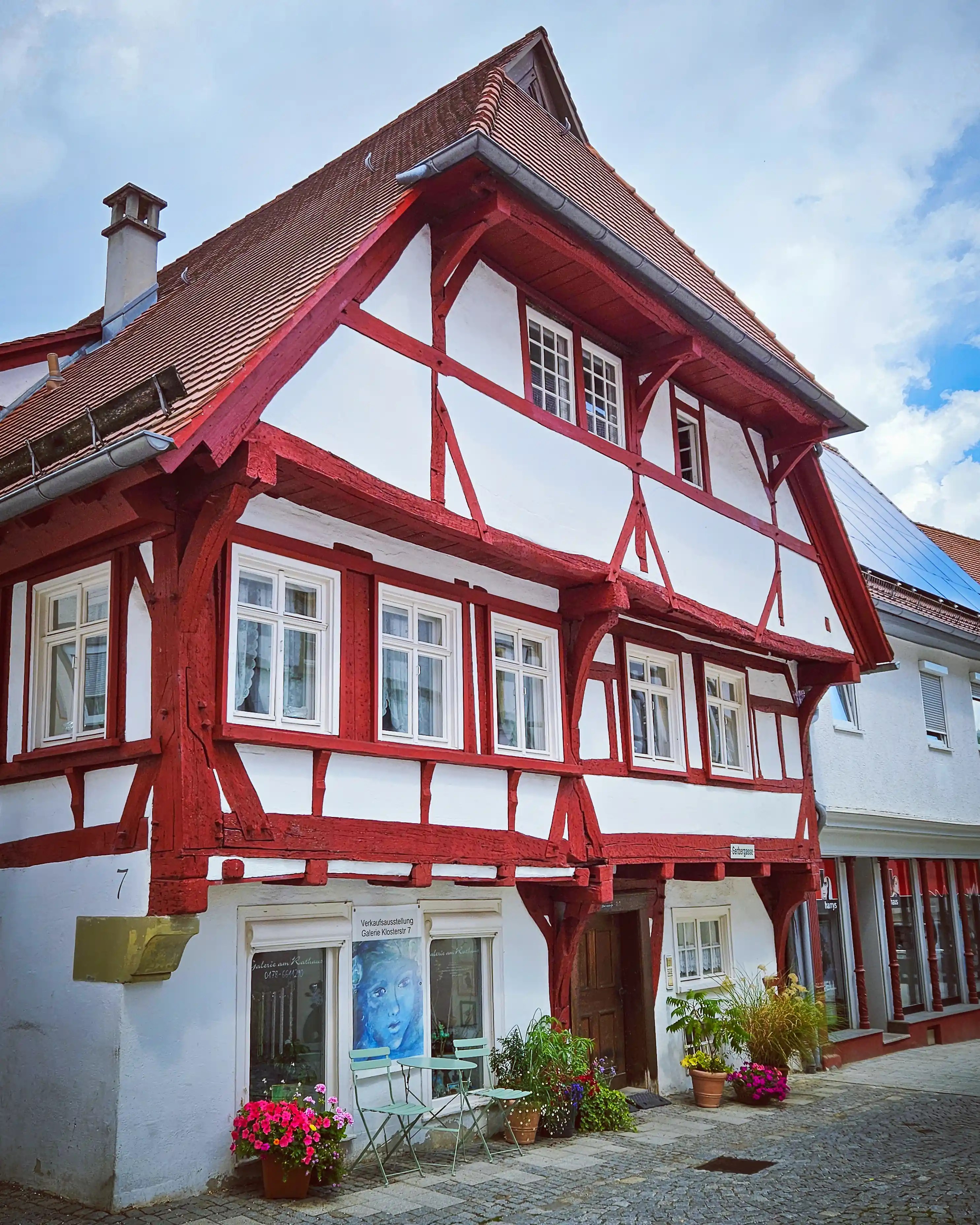 A red and white half-timbered house with flower pots stands along a cobblestone street in Blaubeuren.