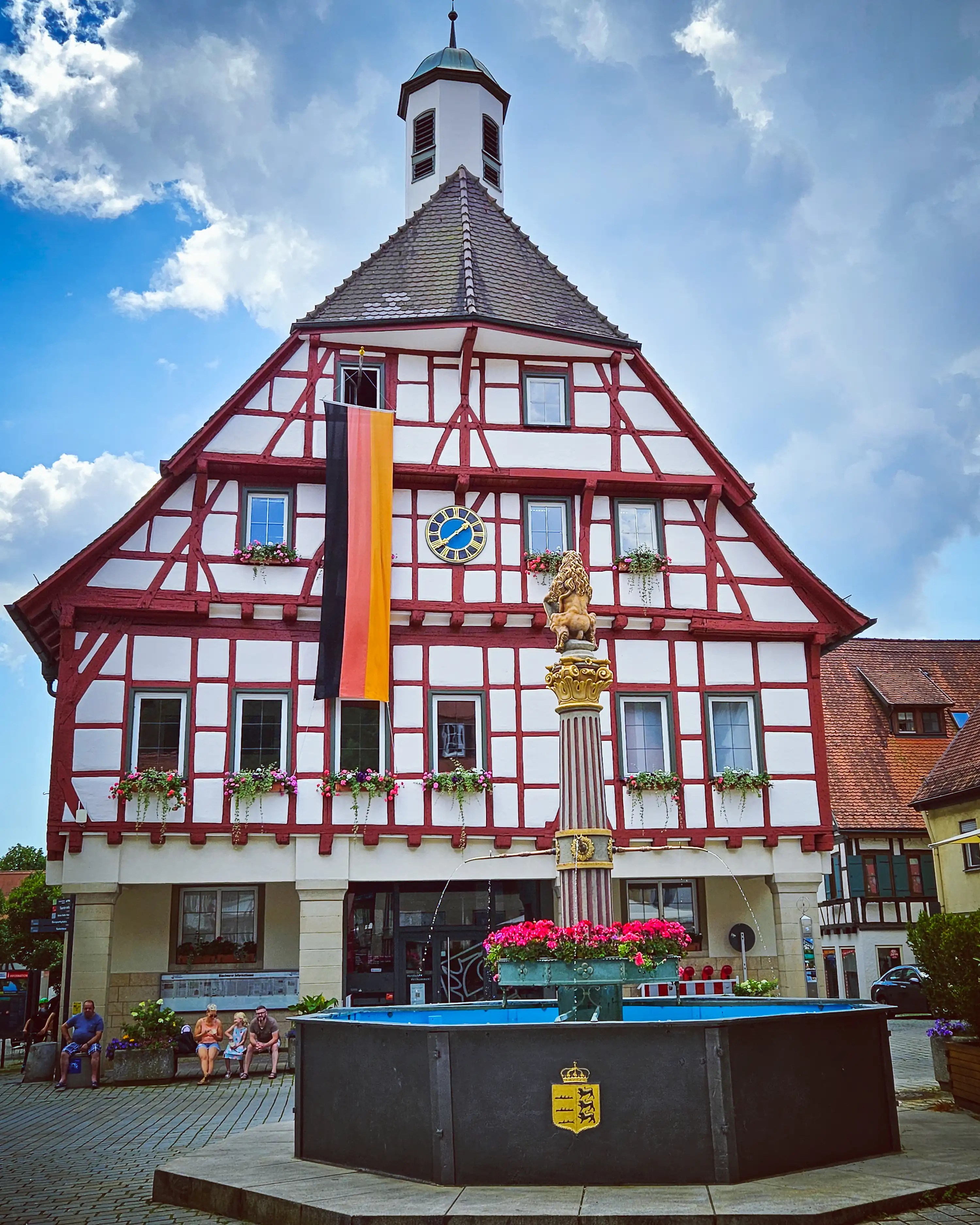 Blaubeuren’s town hall stands behind a fountain with flowers, decorated with a German flag hanging from its façade.