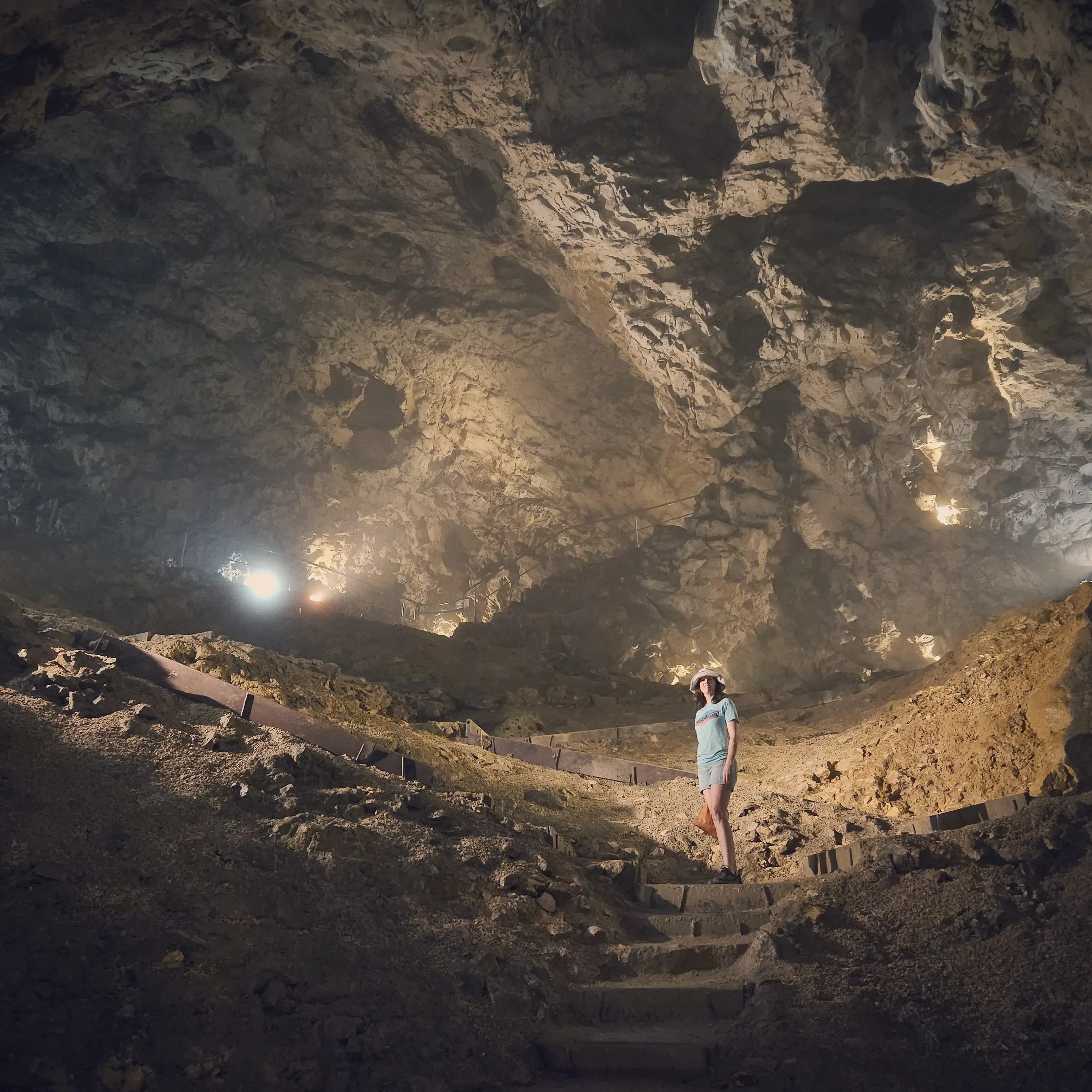 A person stands on stone steps inside a vast cavern chamber at Hohle Fels Cave, surrounded by towering rock walls lit by soft lights.