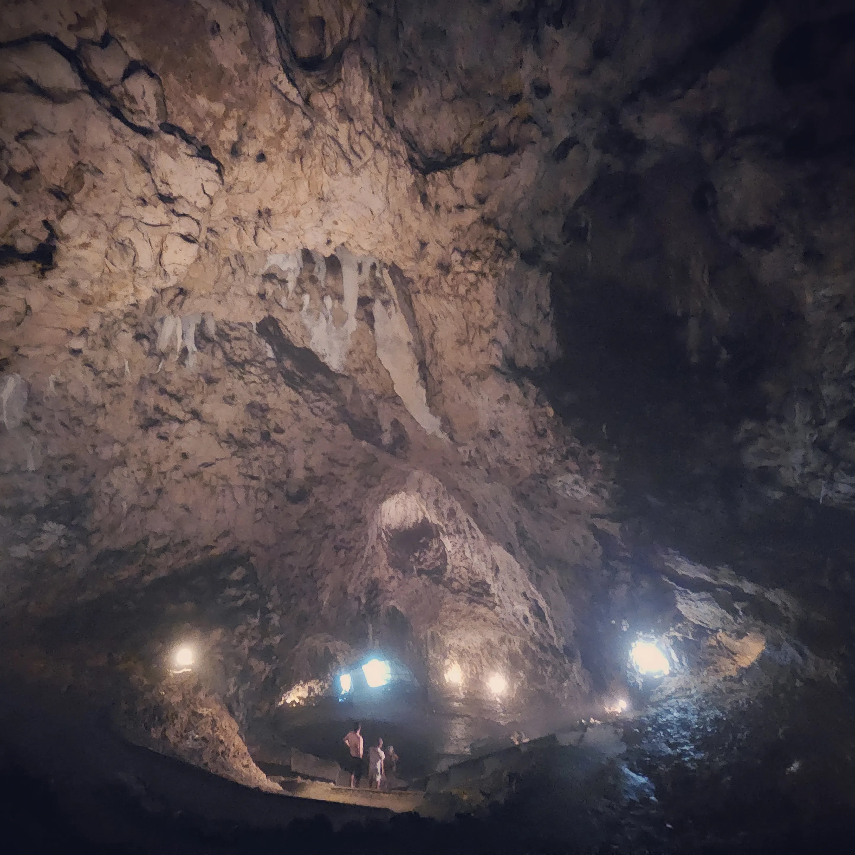 Visitors stand on a walkway inside Hohle Fels Cave beneath a high ceiling of rugged limestone illuminated by warm lights.