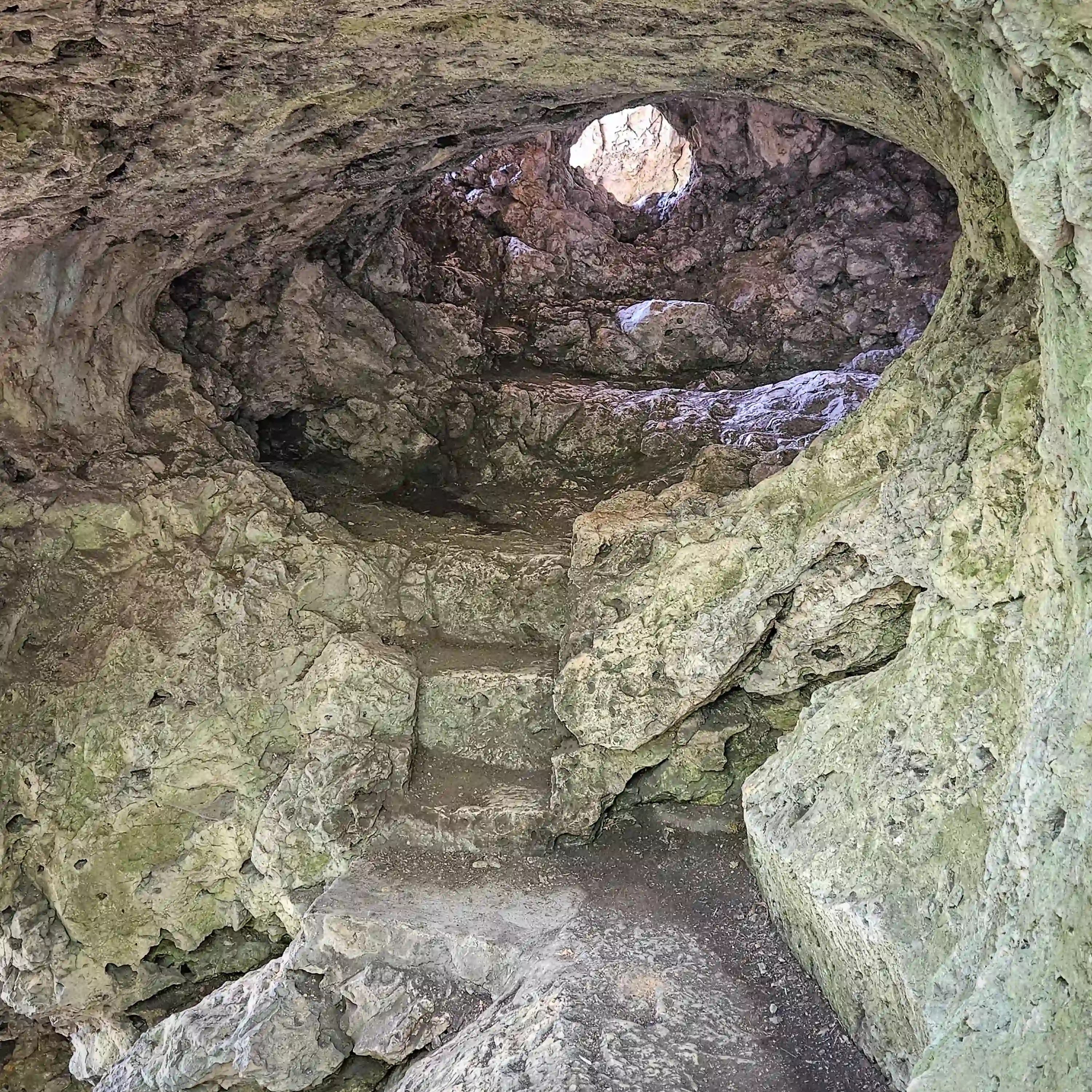 Rough stone steps carved into natural rock lead upward inside a small cave opening with daylight visible through a round hole in the rock.