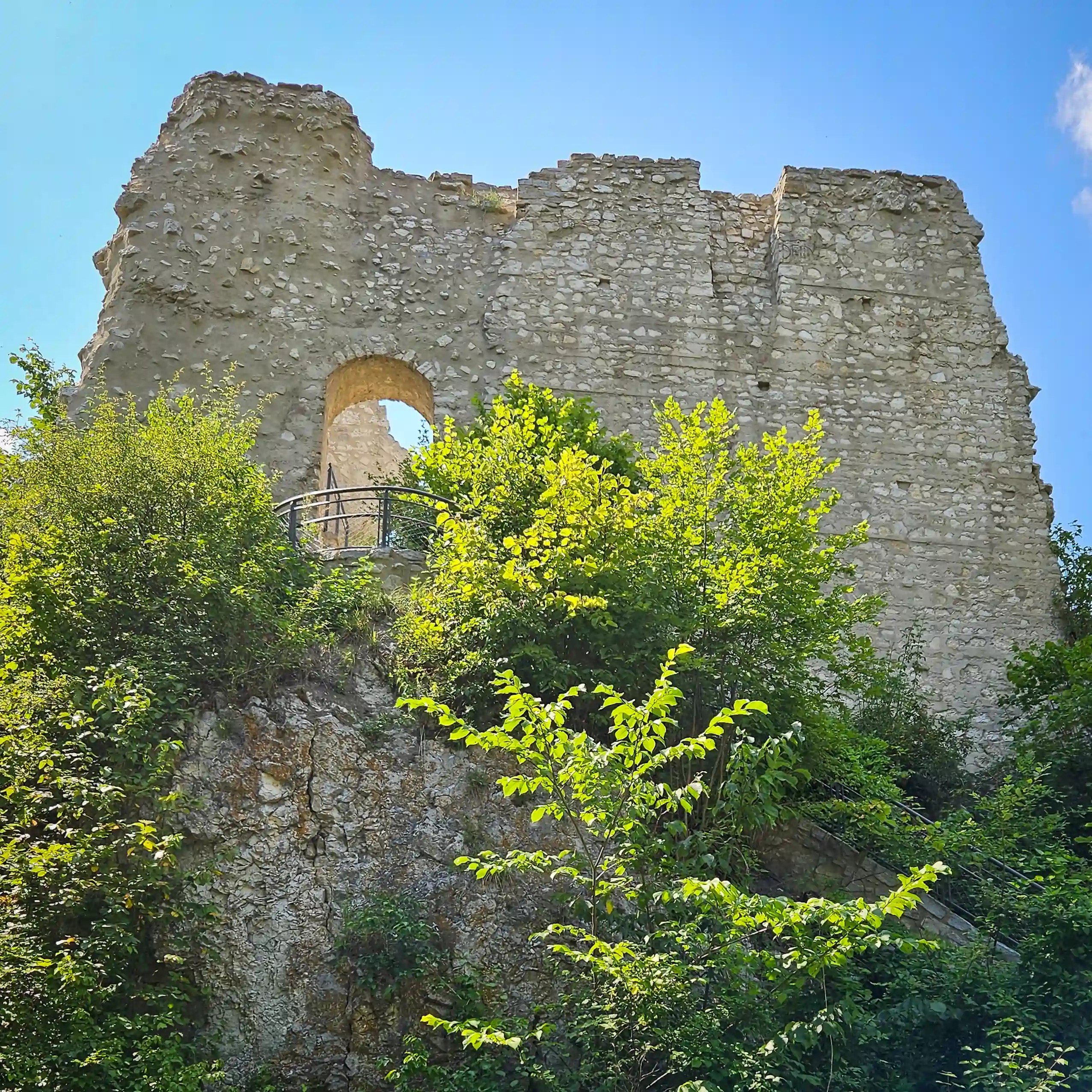 A large stone wall with an arched opening stands on a rocky hillside surrounded by trees under a bright blue sky.