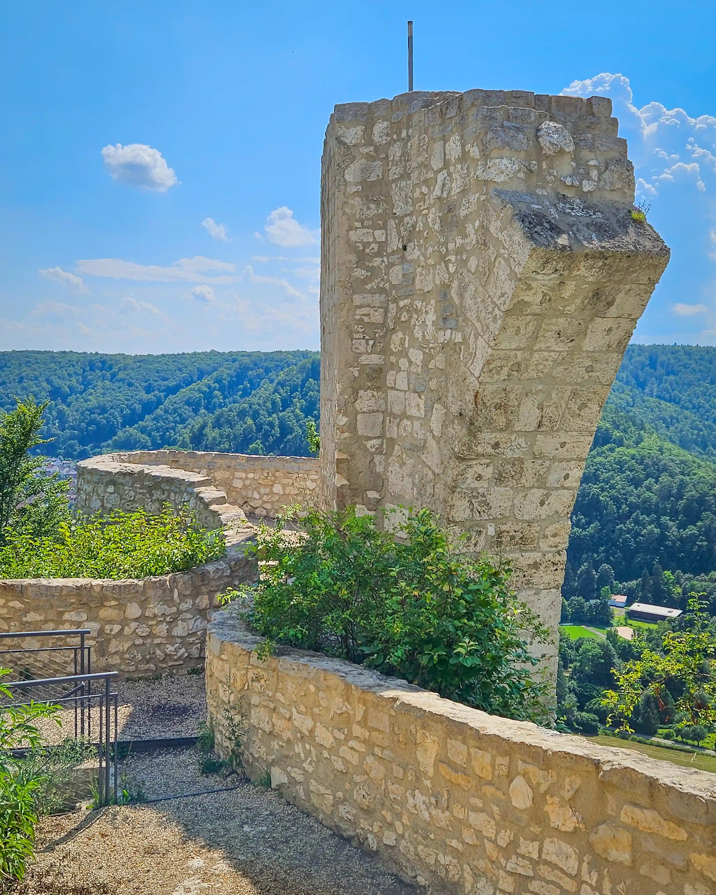 A tall rectangular stone tower remnant stands above a curved defensive wall, overlooking a green valley under a blue sky with scattered clouds.