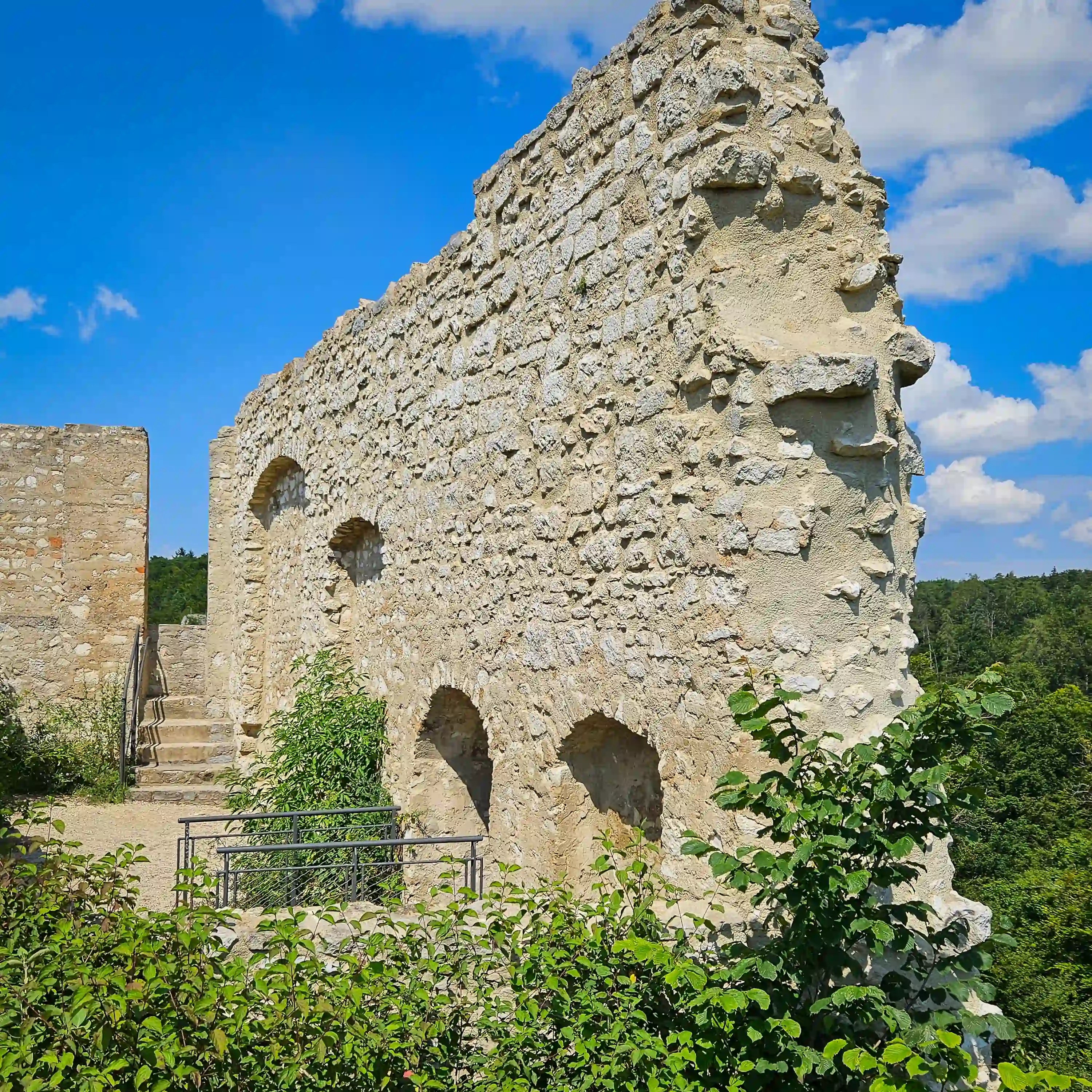 A thick stone wall with several small arched openings stands against a bright blue sky, surrounded by green shrubs.