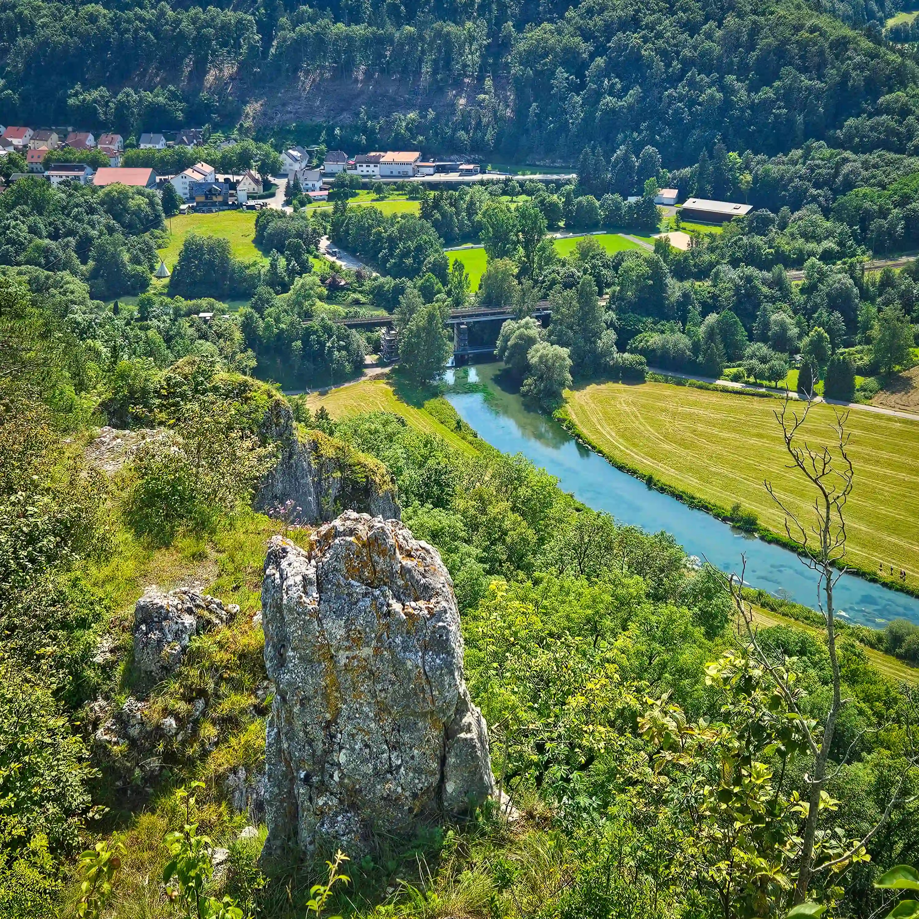 A turquoise river curves through a green valley below rocky cliffs, with houses and buildings visible in the distance.