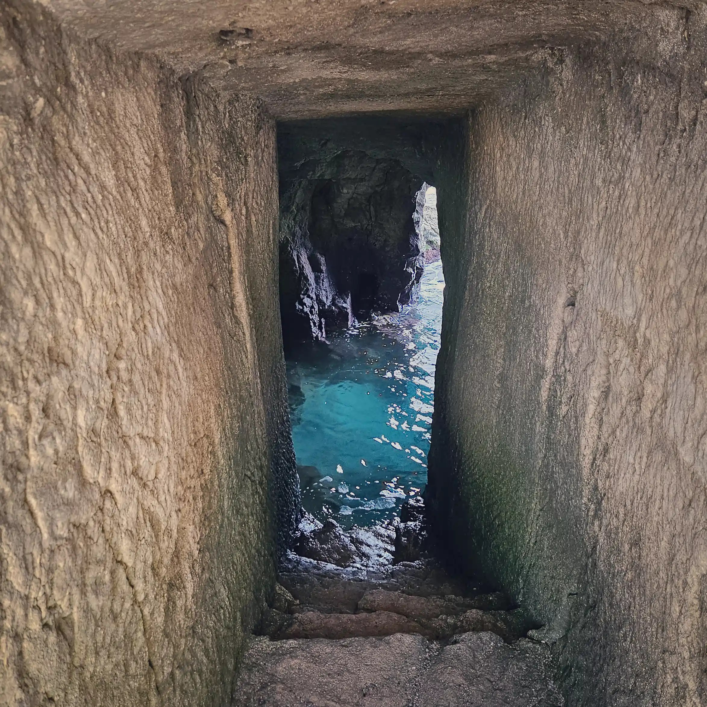 A narrow stone passage leading down to turquoise water inside a small coastal cave.